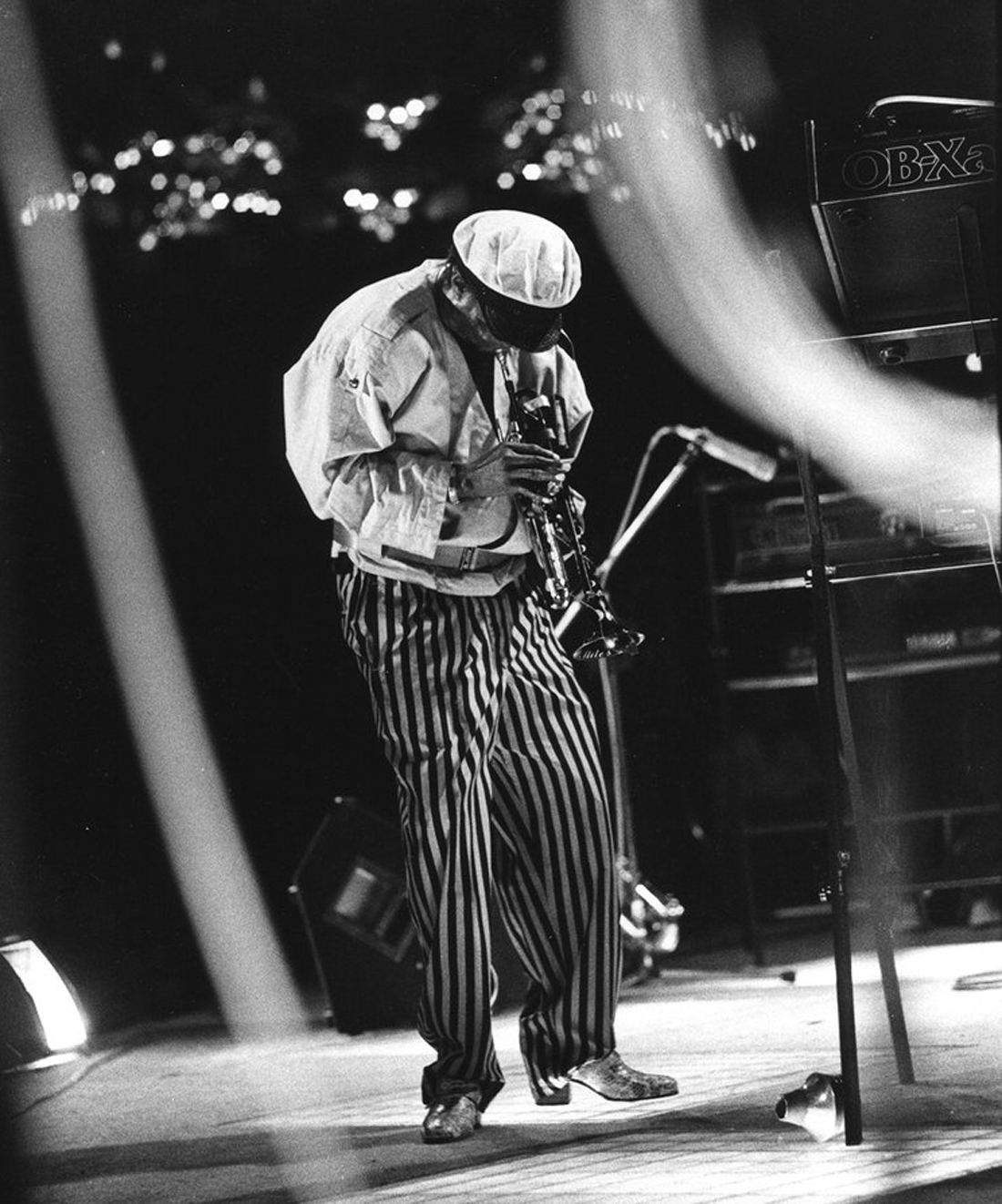 A musician in striped pants and a cap plays the trumpet on stage under dramatic lighting.
