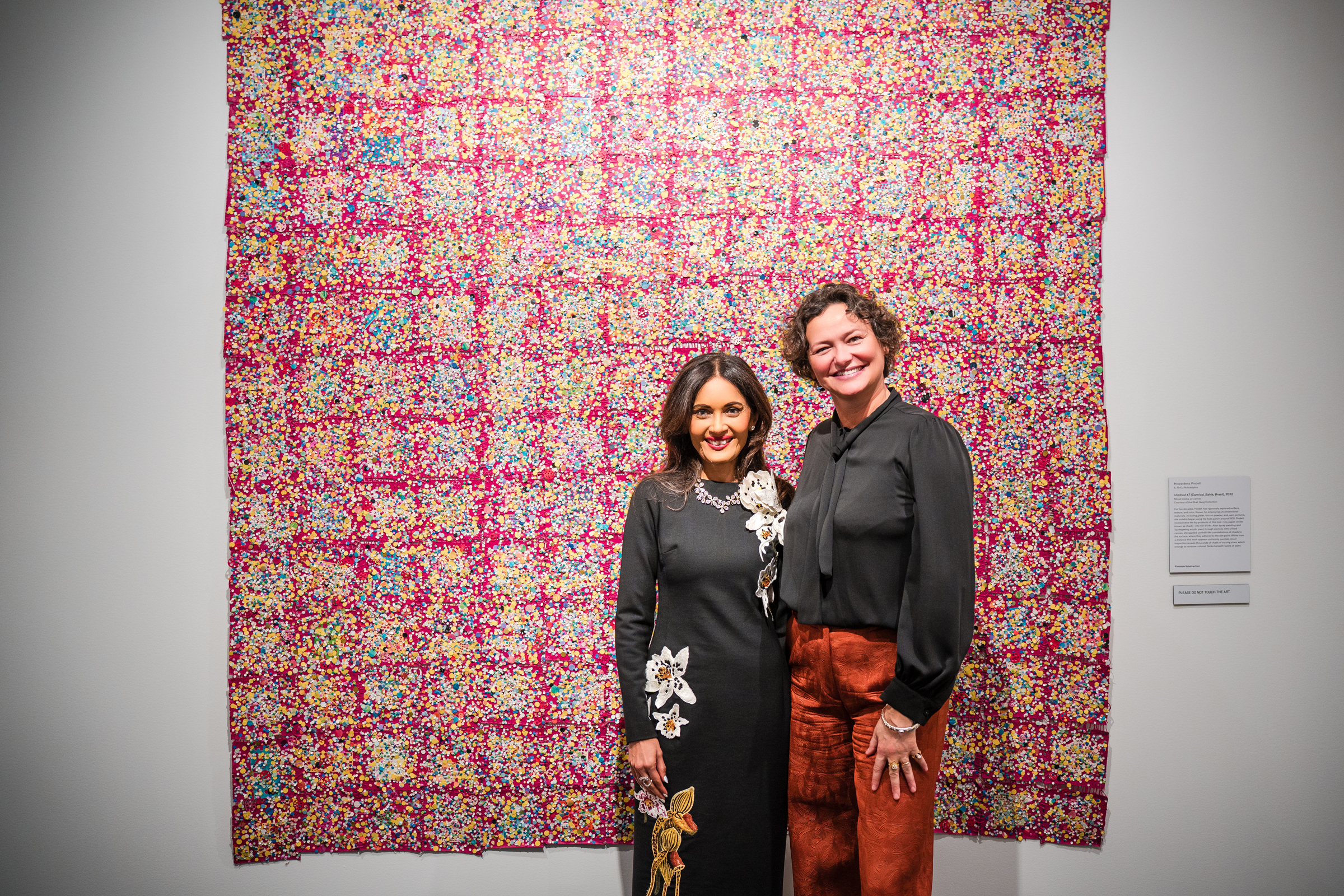 Two people stand in front of a colorful, patterned textile displayed on a gallery wall.