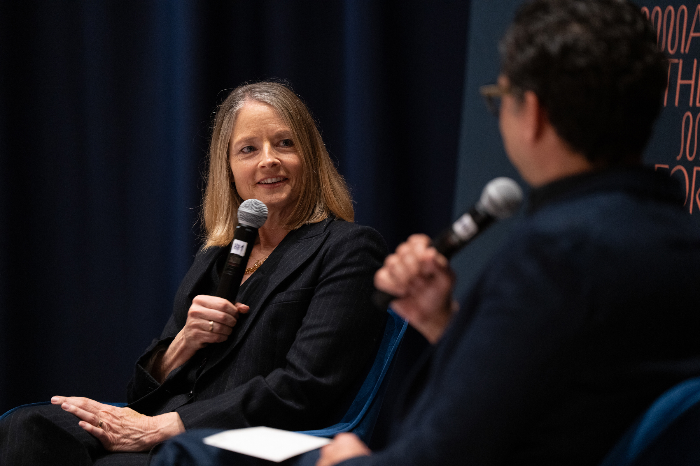 Two people are sitting and speaking into microphones during an event or discussion.
