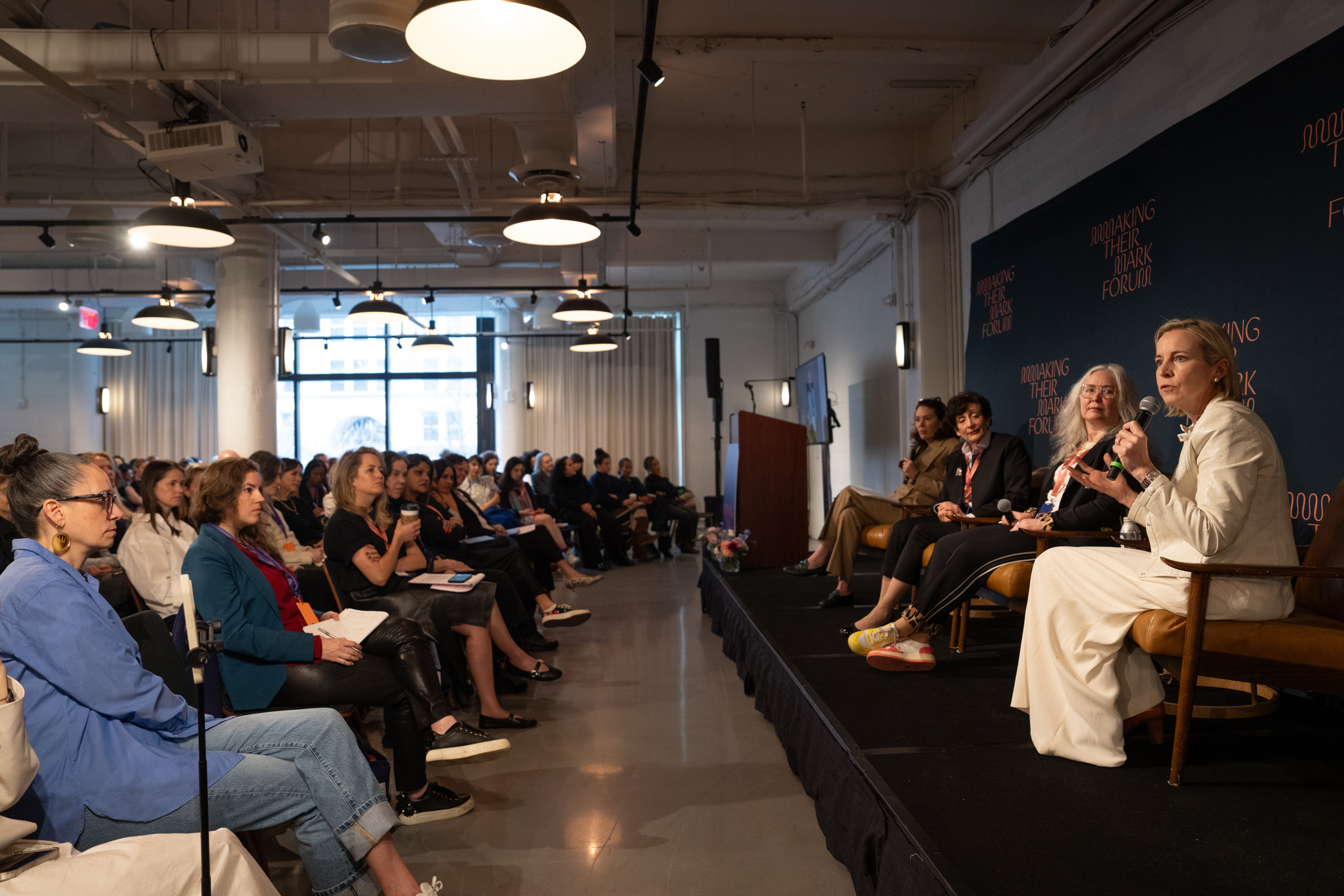 A panel of speakers sits on stage addressing an audience at an indoor forum event.