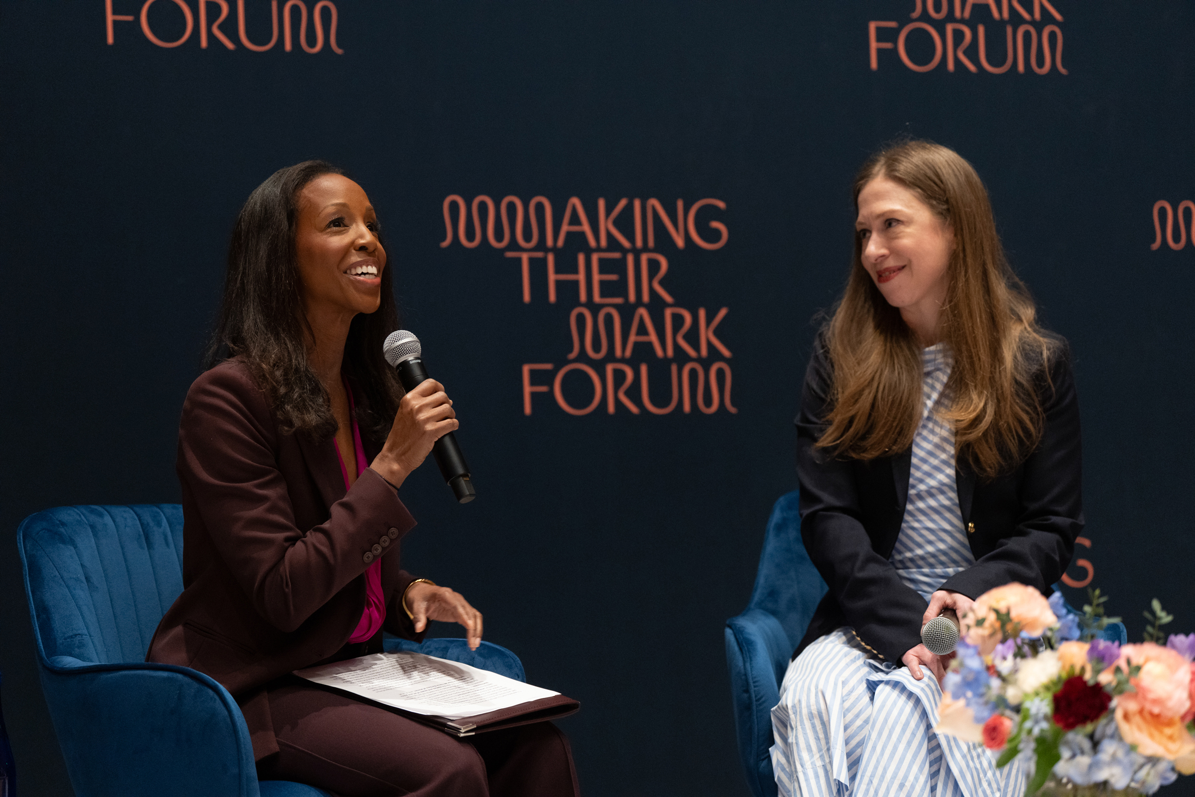 Two people are seated on stage in conversation, holding microphones at a forum event.