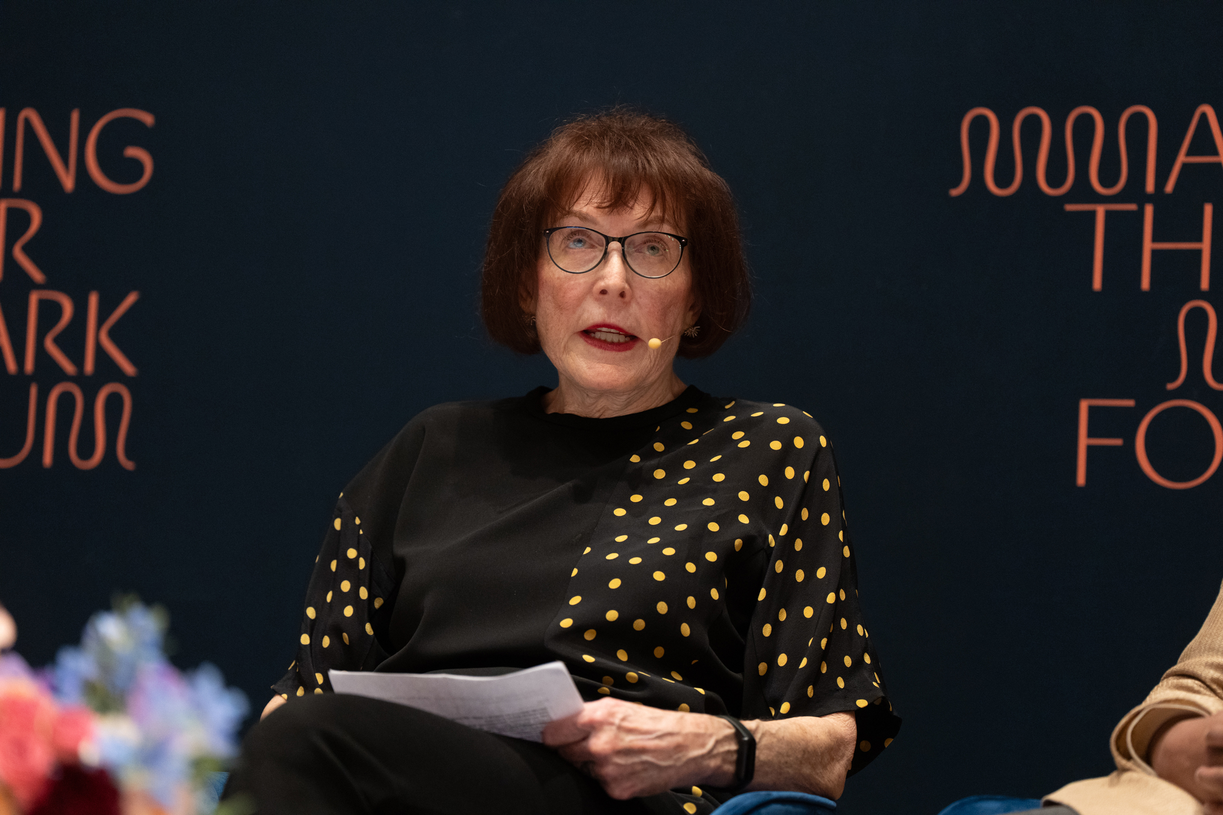 A person is sitting and holding papers, wearing a black shirt with yellow polka dots.