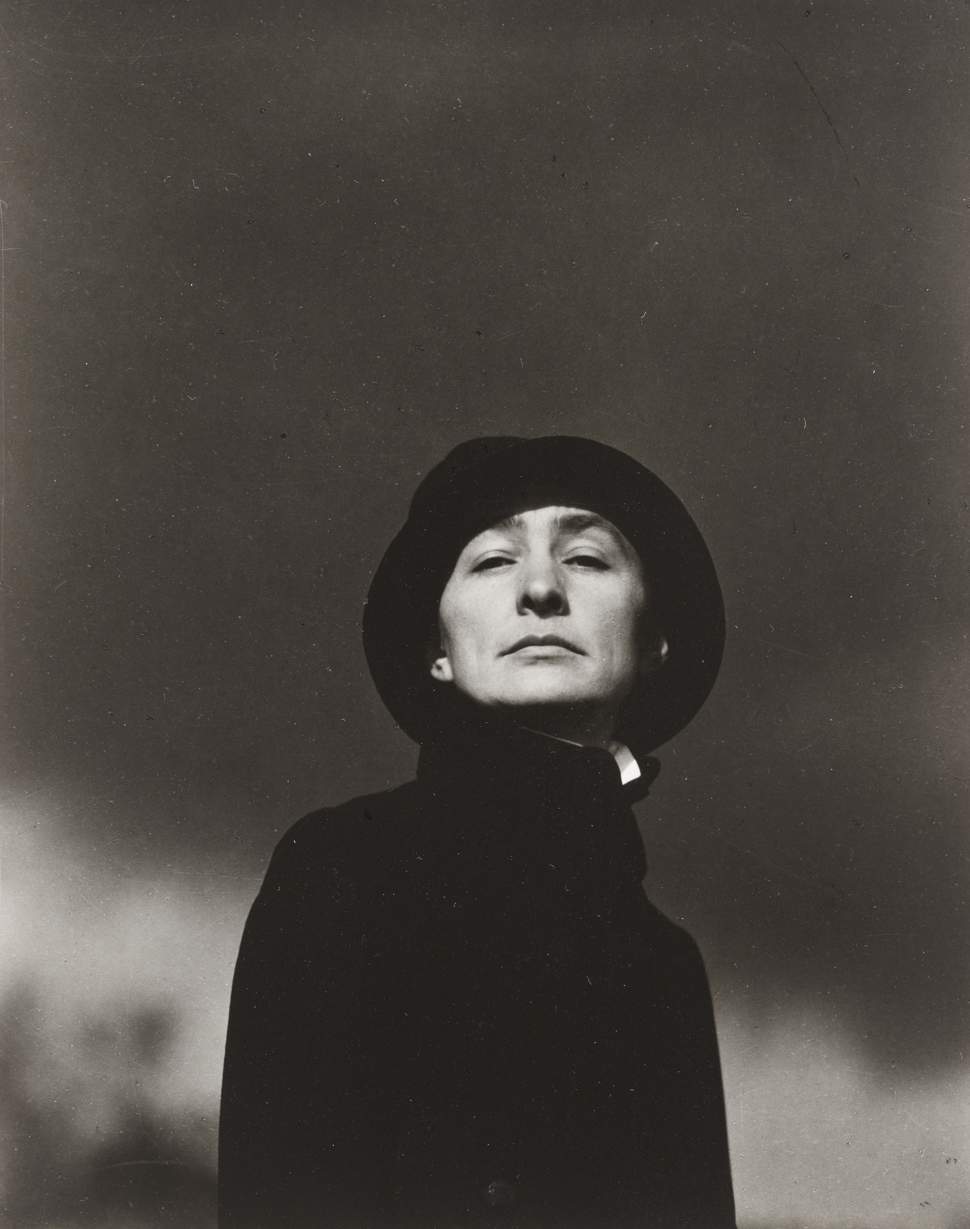A person in a dark coat and hat stands against a moody, overcast background.