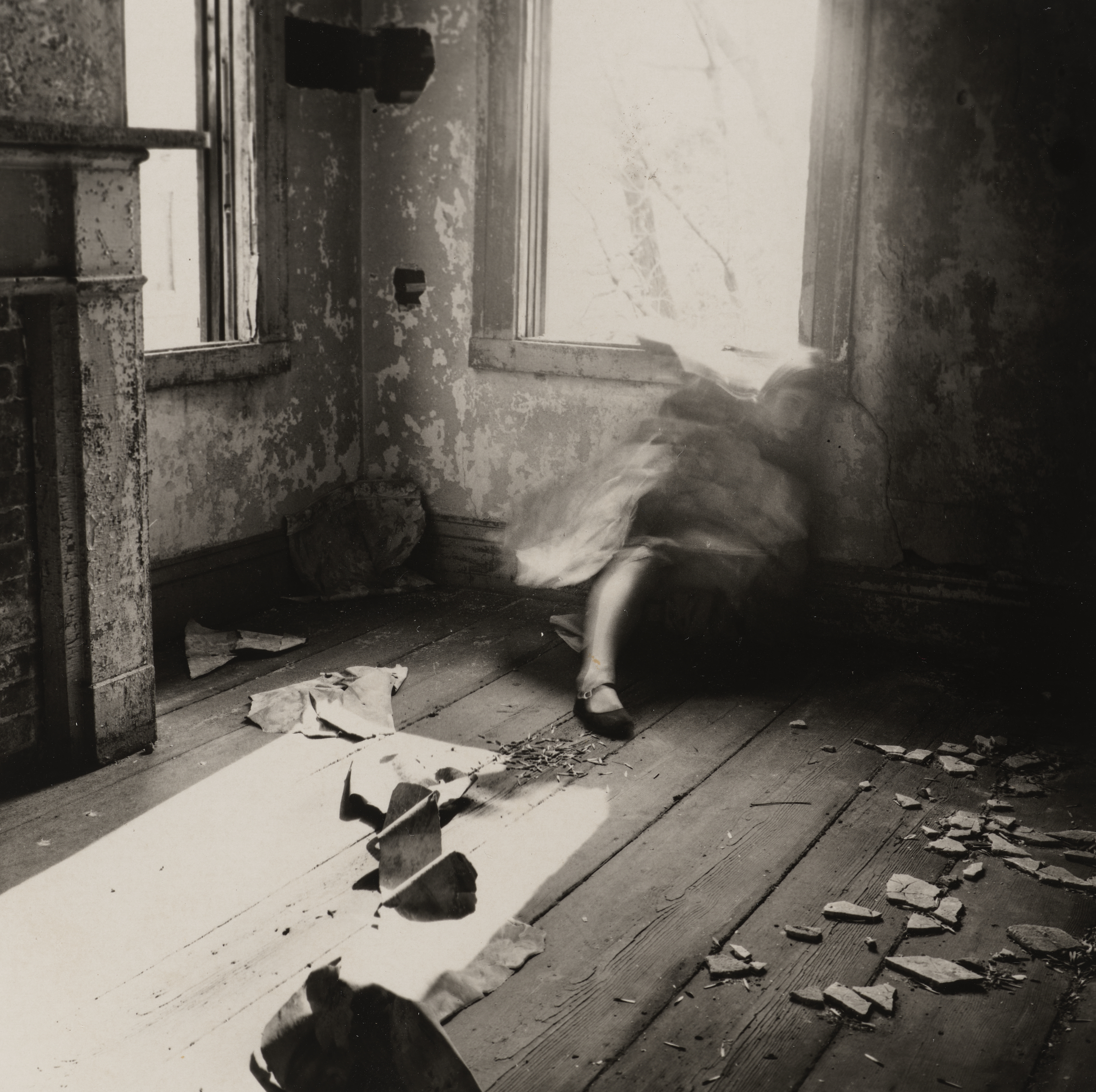 A blurred figure sits against the wall in a decaying, sunlit room with broken debris on the floor.