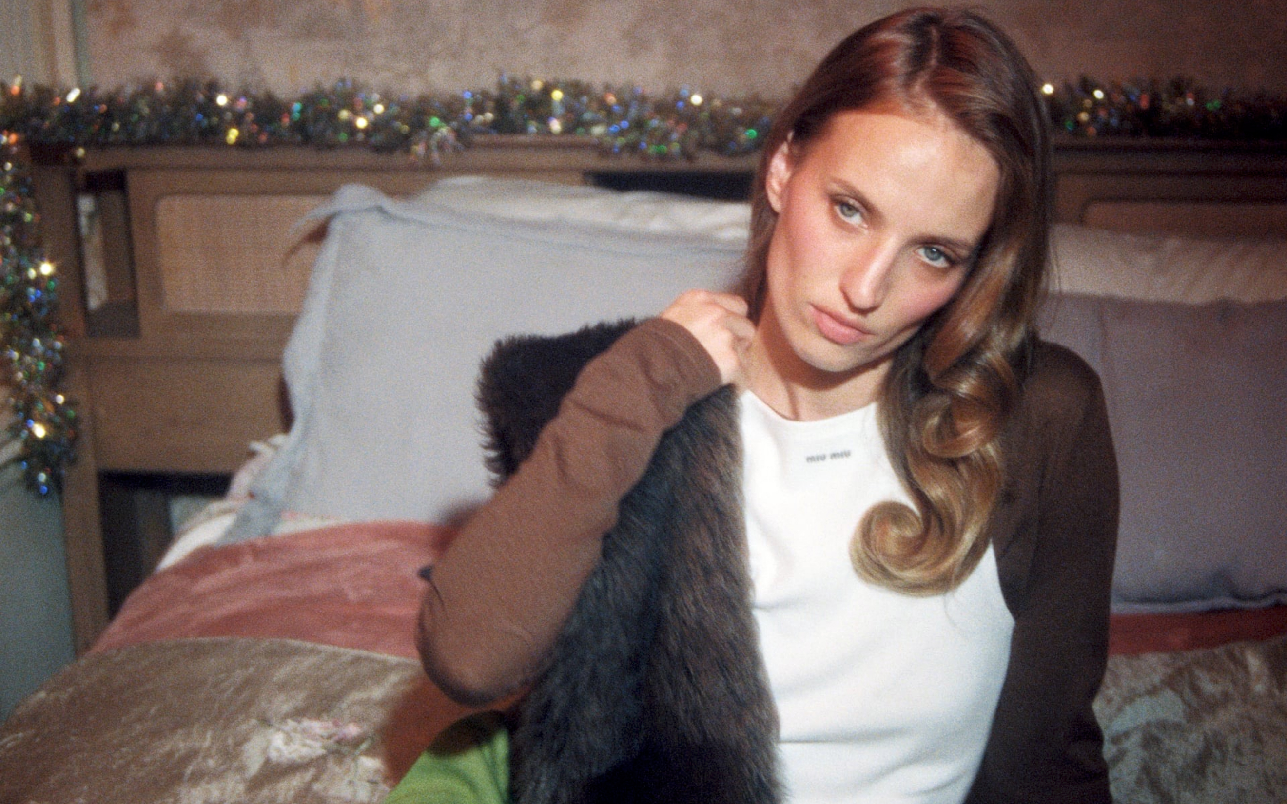 A person with long hair sits on a bed with festive garland decorations behind them.