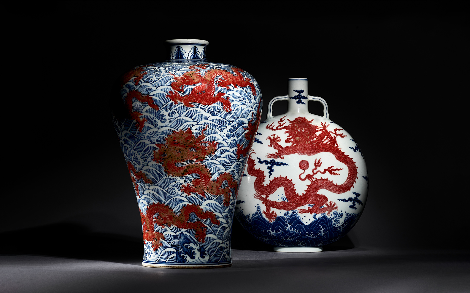 Two ornate vases with red dragons and blue waves are displayed against a dark background. | Two ornate Chinese porcelain vases feature red dragons and blue waves on a dark background.