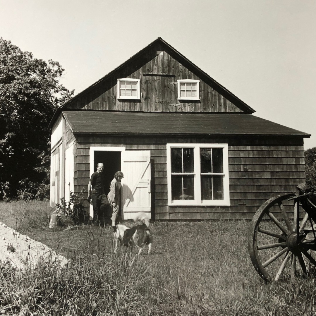 Two people and a dog stand outside a rustic wooden house with a wagon wheel in the foreground.
