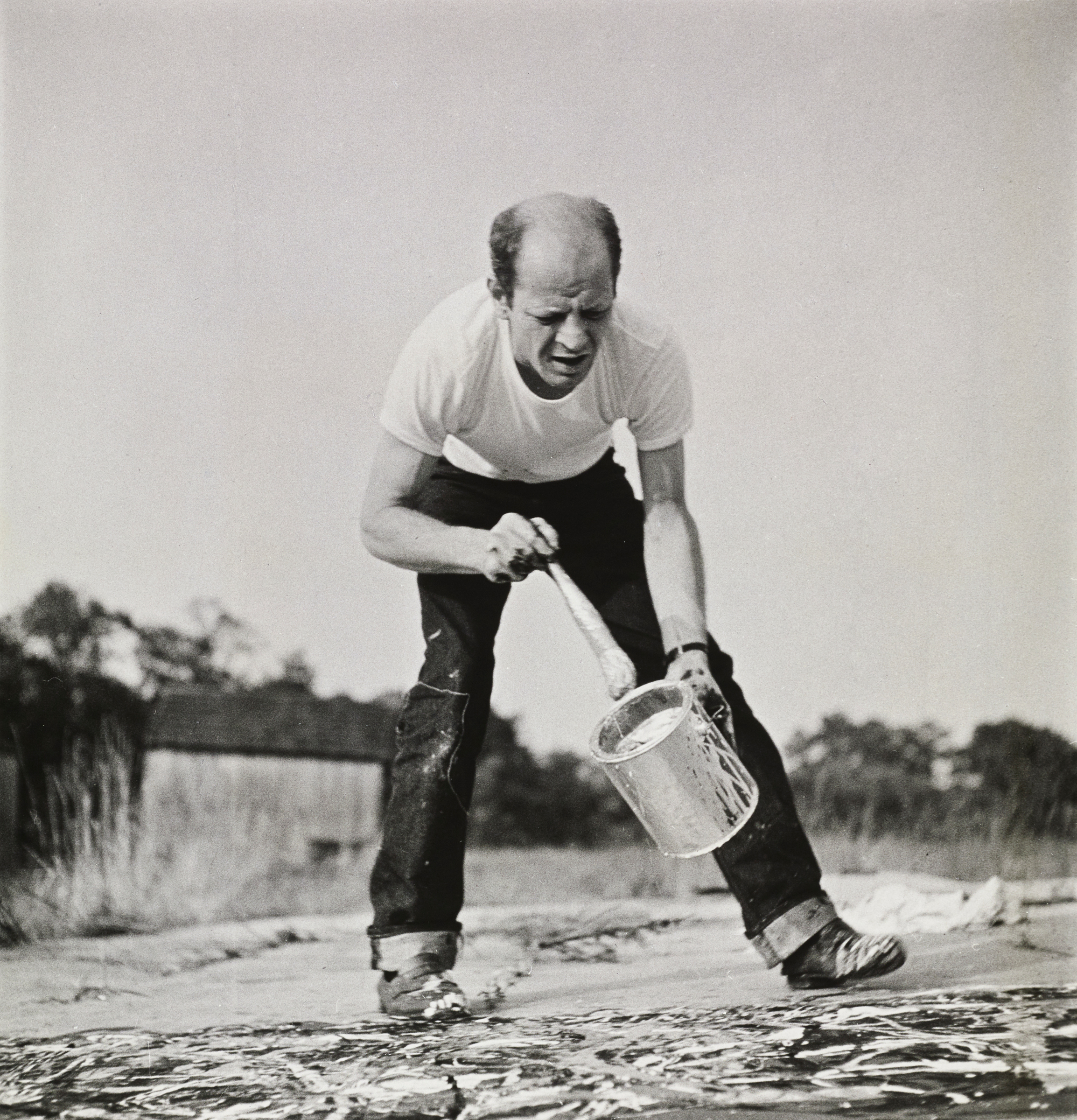 A person in a white shirt pours paint onto a canvas outdoors using a large paint can.