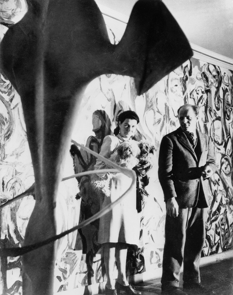 Two people stand in front of an abstract mural, with one holding a dog and a sculpture in the foreground.