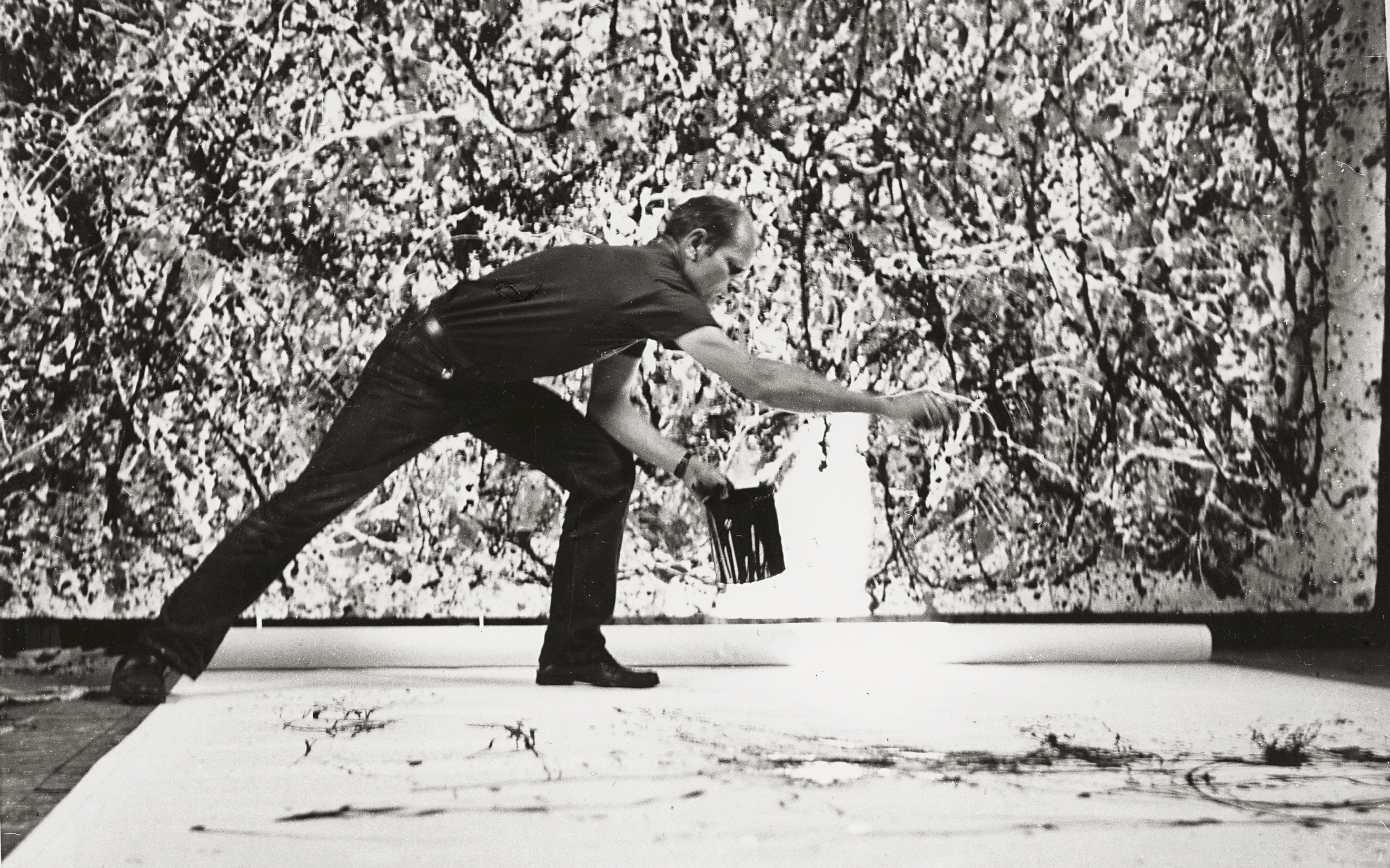 A person is painting on a large canvas using the drip technique in a studio.