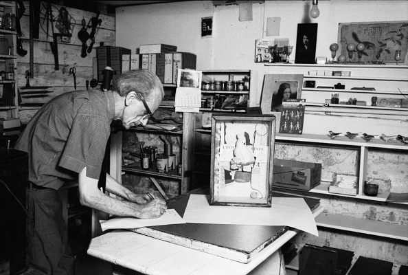 An elderly man works at a table in a cluttered art studio filled with books and tools.