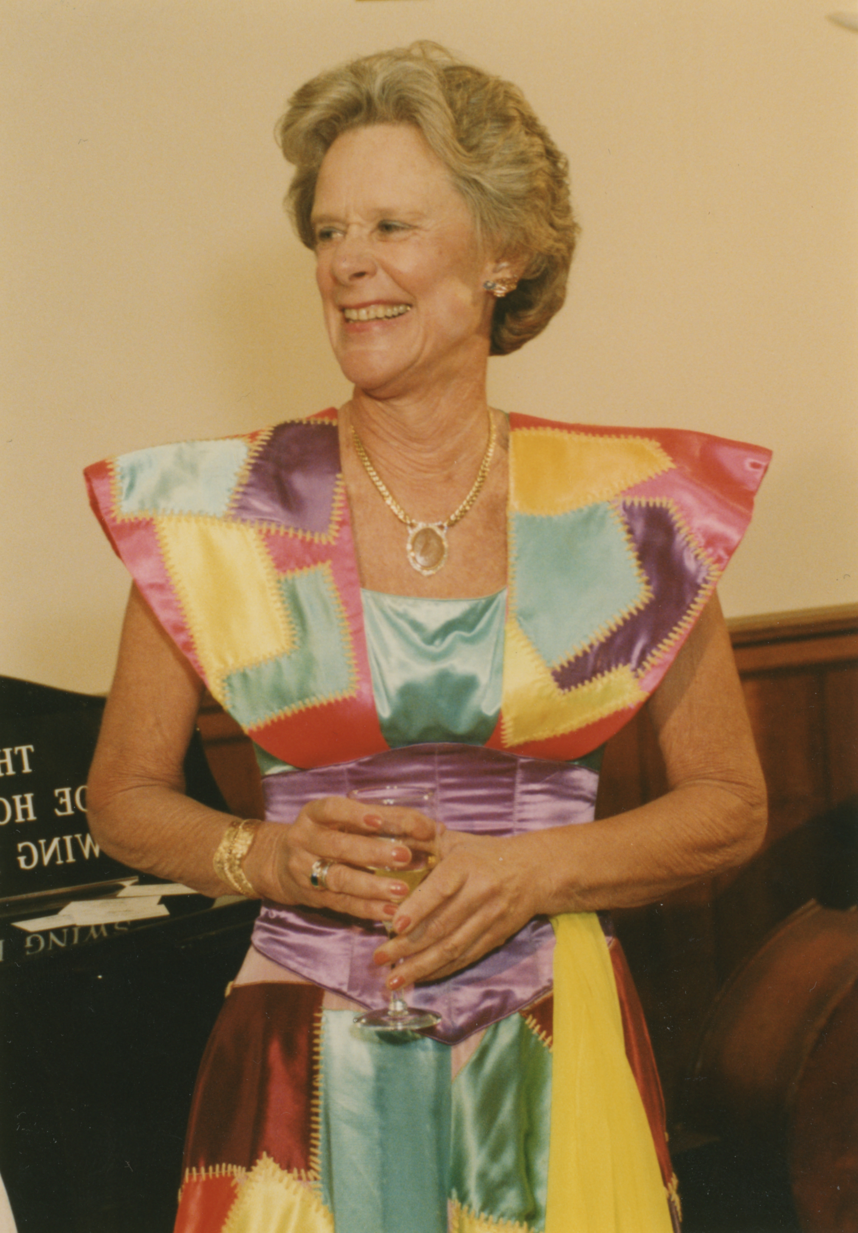A woman in a colorful patchwork dress holds a glass and wears gold jewelry.