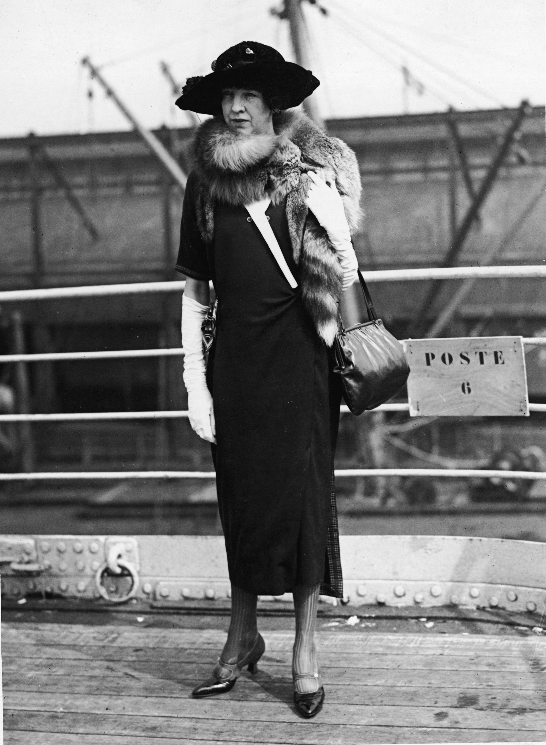 A woman in vintage attire stands on a ship deck wearing a fur stole and hat.