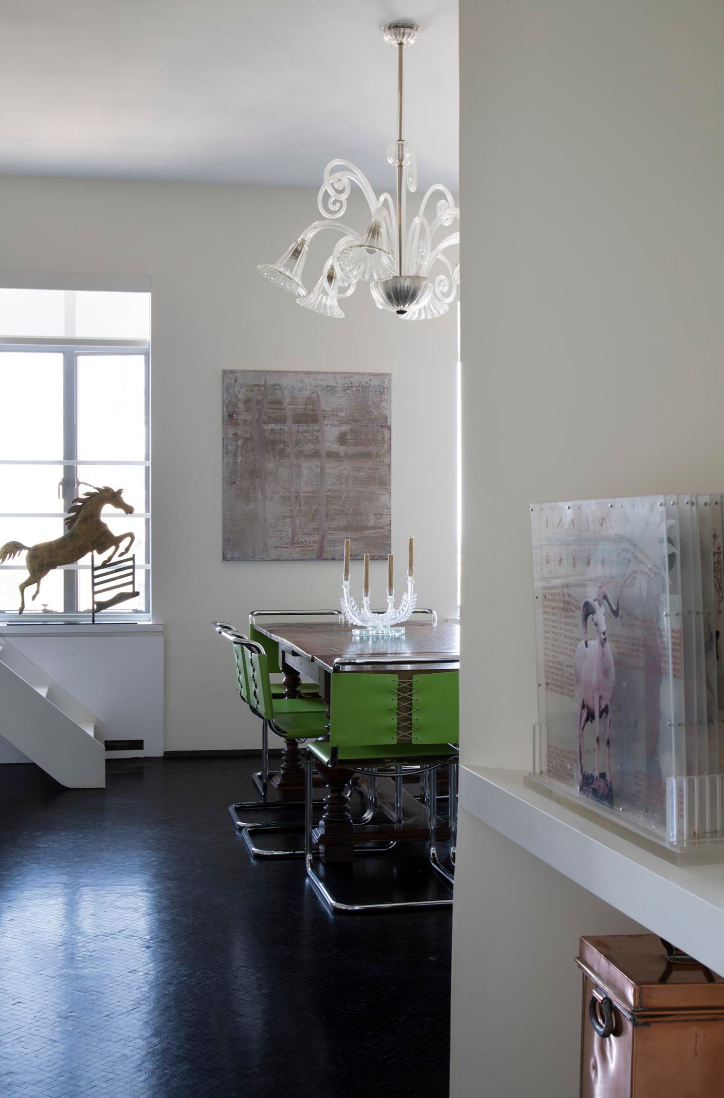A dining room with green chairs, a modern chandelier, art, and a horse sculpture by the window.