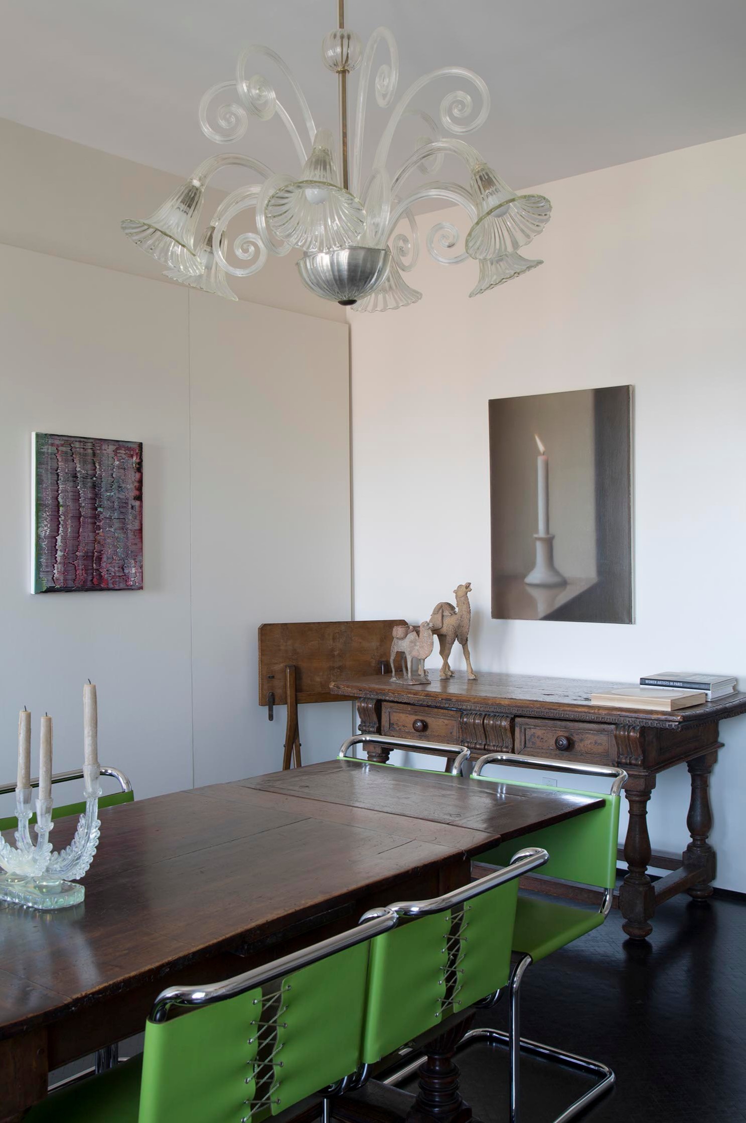 A dining room with a wooden table, green chairs, a chandelier, and eclectic decor on the wall.