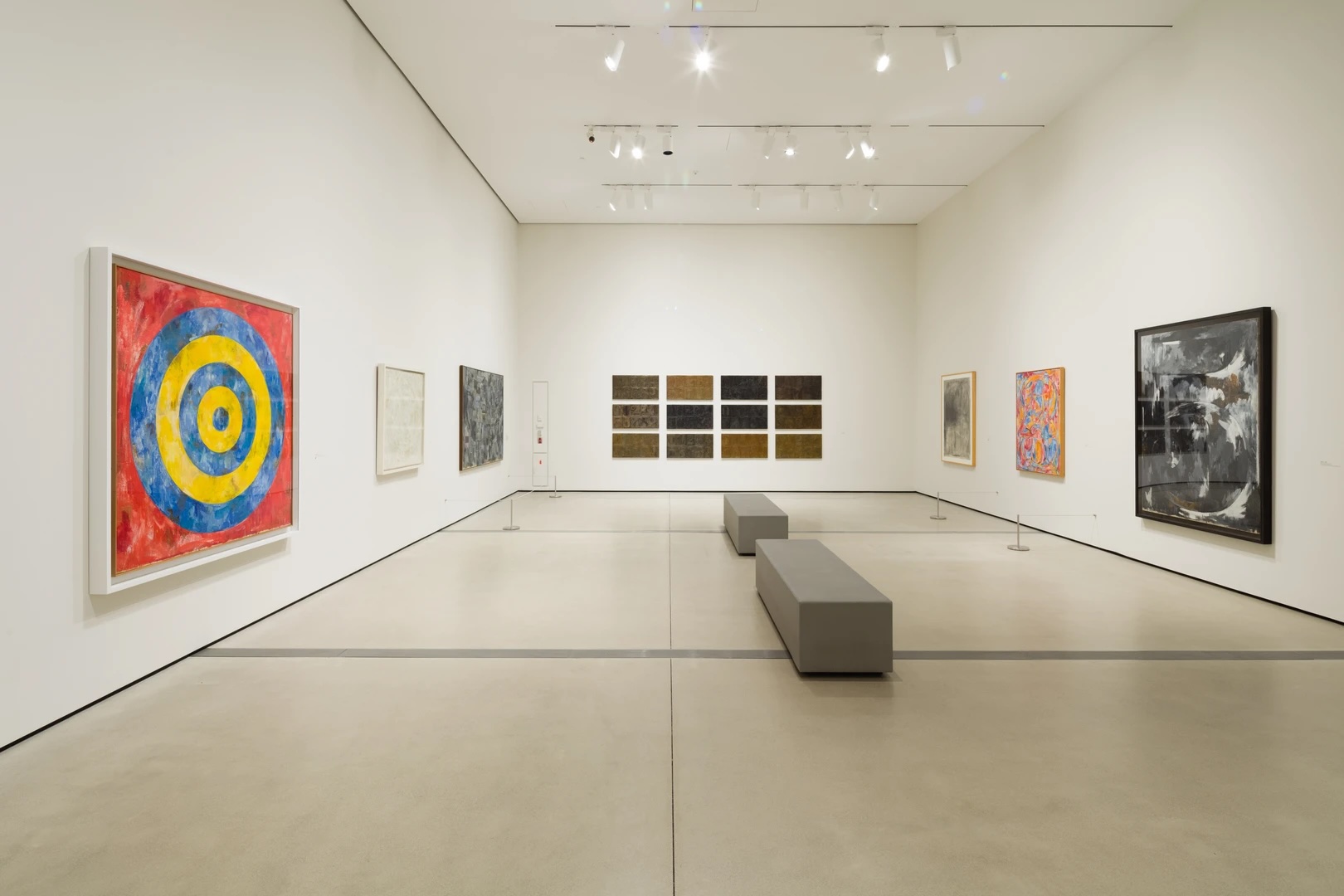 A modern art gallery with abstract paintings on white walls and two gray benches in the center.