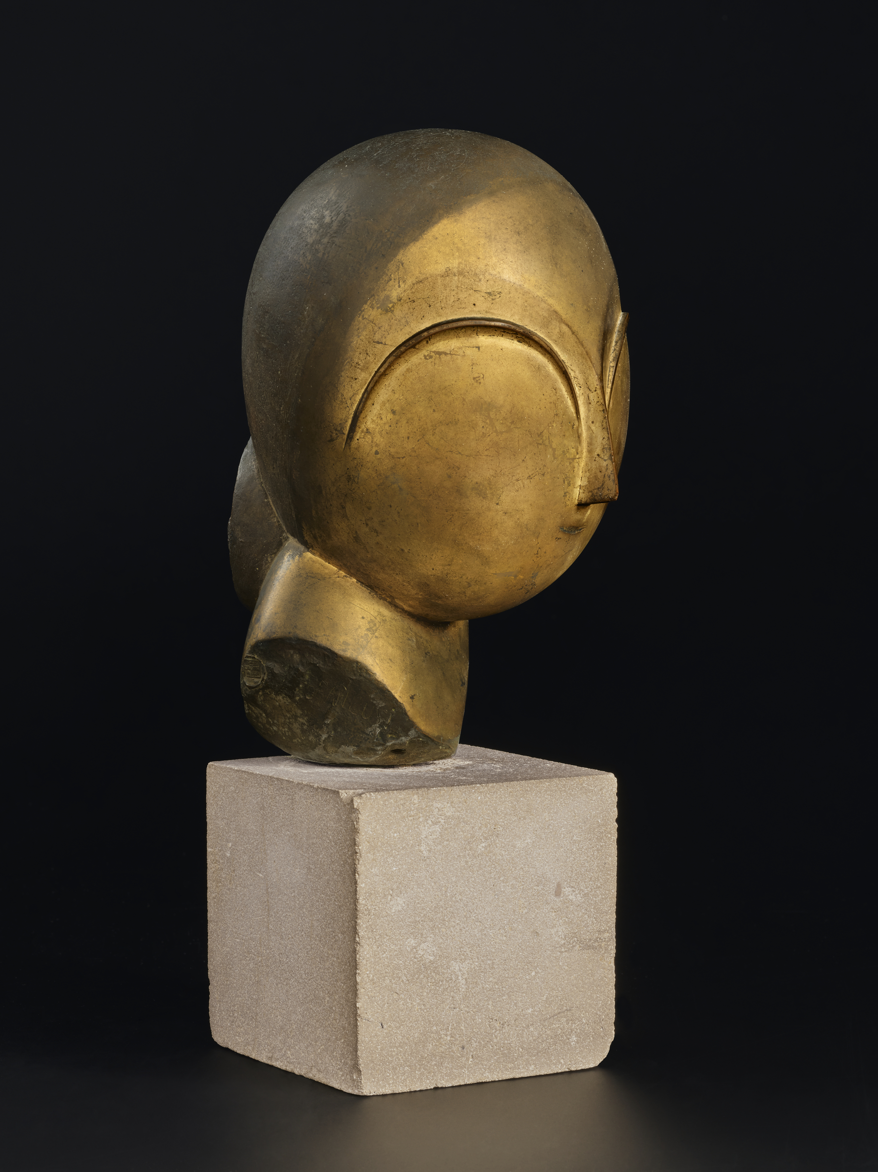 A smooth, abstract golden head sculpture sits atop a square stone pedestal against a dark background.
