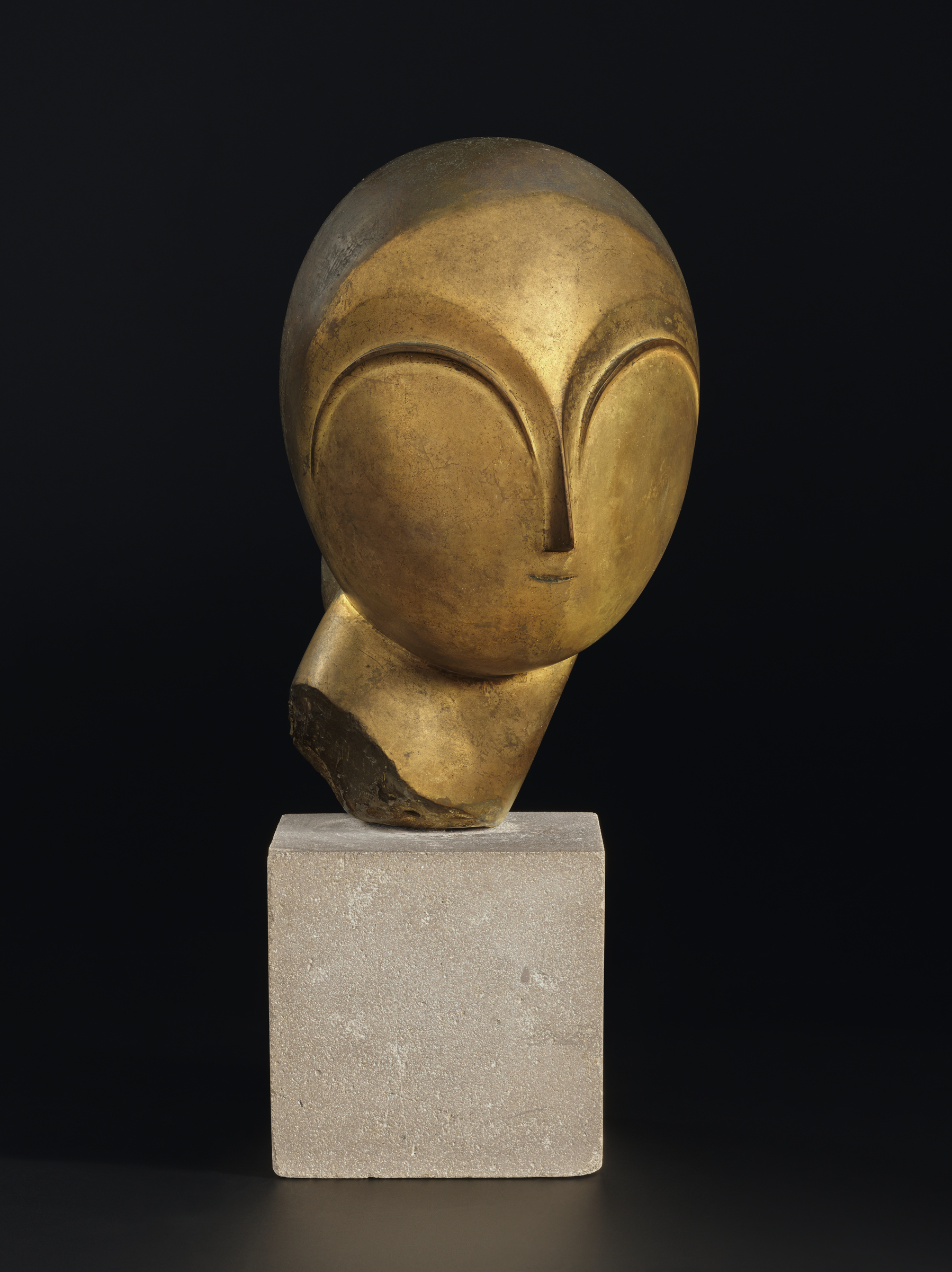 A golden abstract sculpture of a head with minimal facial features sits on a stone pedestal.