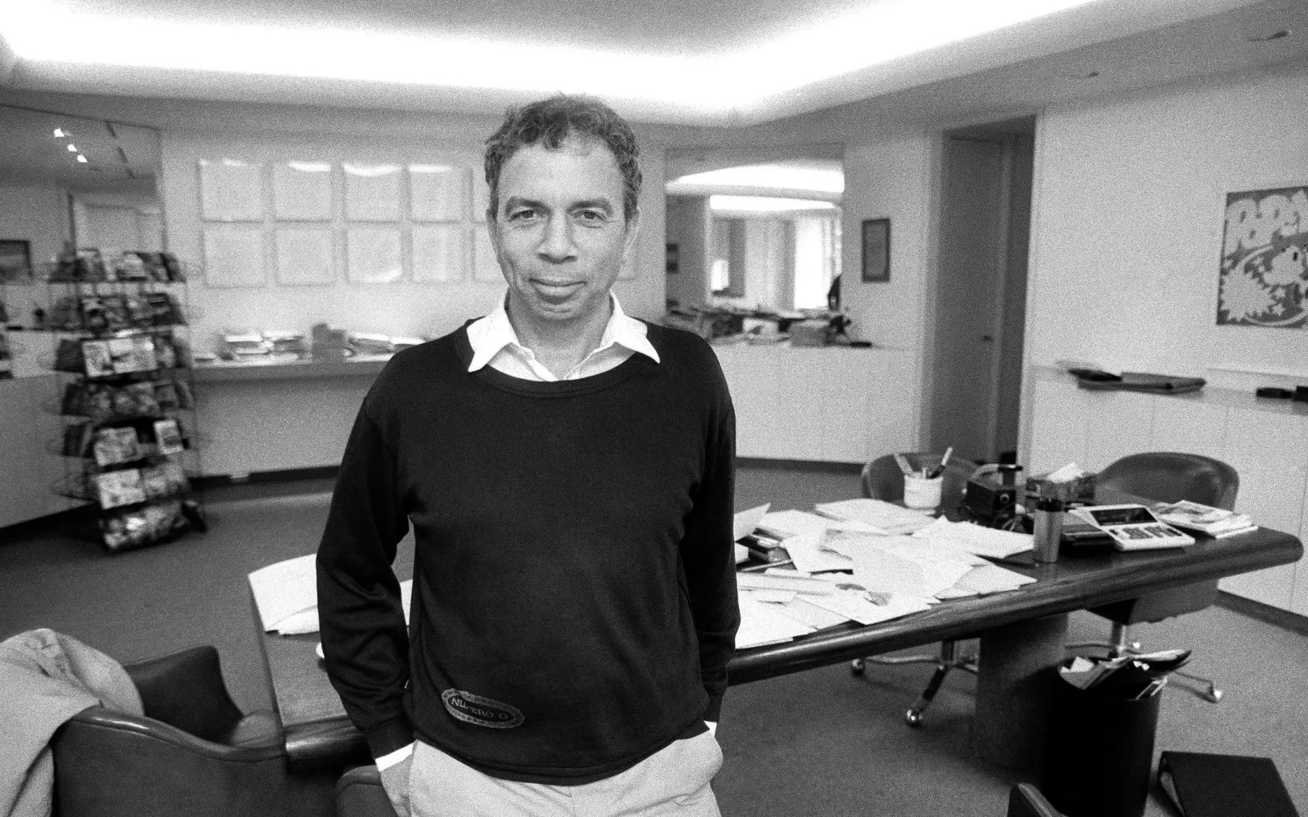A person stands in a modern office with a cluttered desk and shelves in the background.