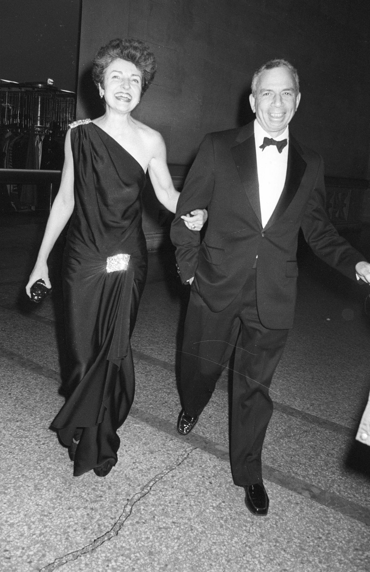 A couple dressed formally walk arm in arm, the woman in a black gown, the man in a tuxedo.