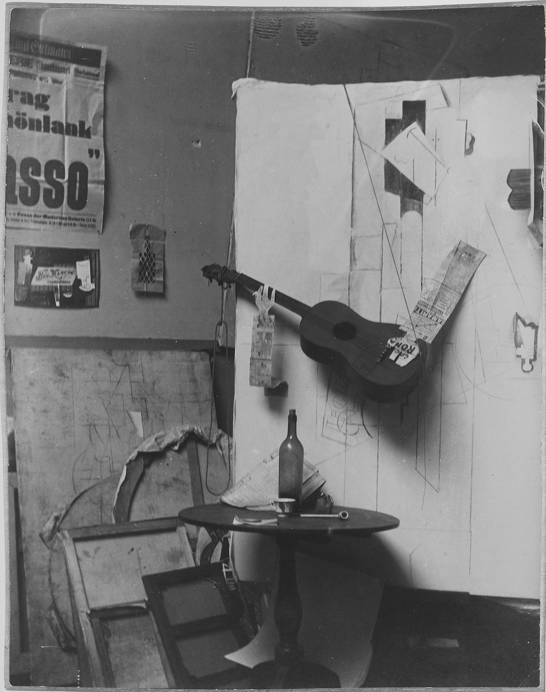 A cluttered artist's studio features a guitar, sketches, posters, and a bottle on a table.