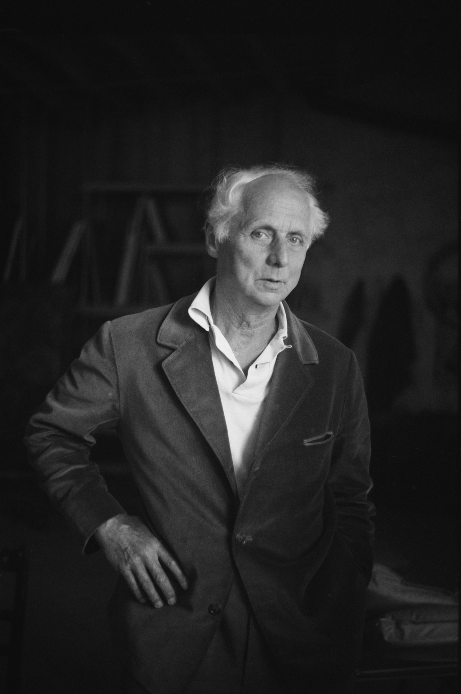 An elderly man with white hair stands indoors wearing a jacket and shirt, face blurred.