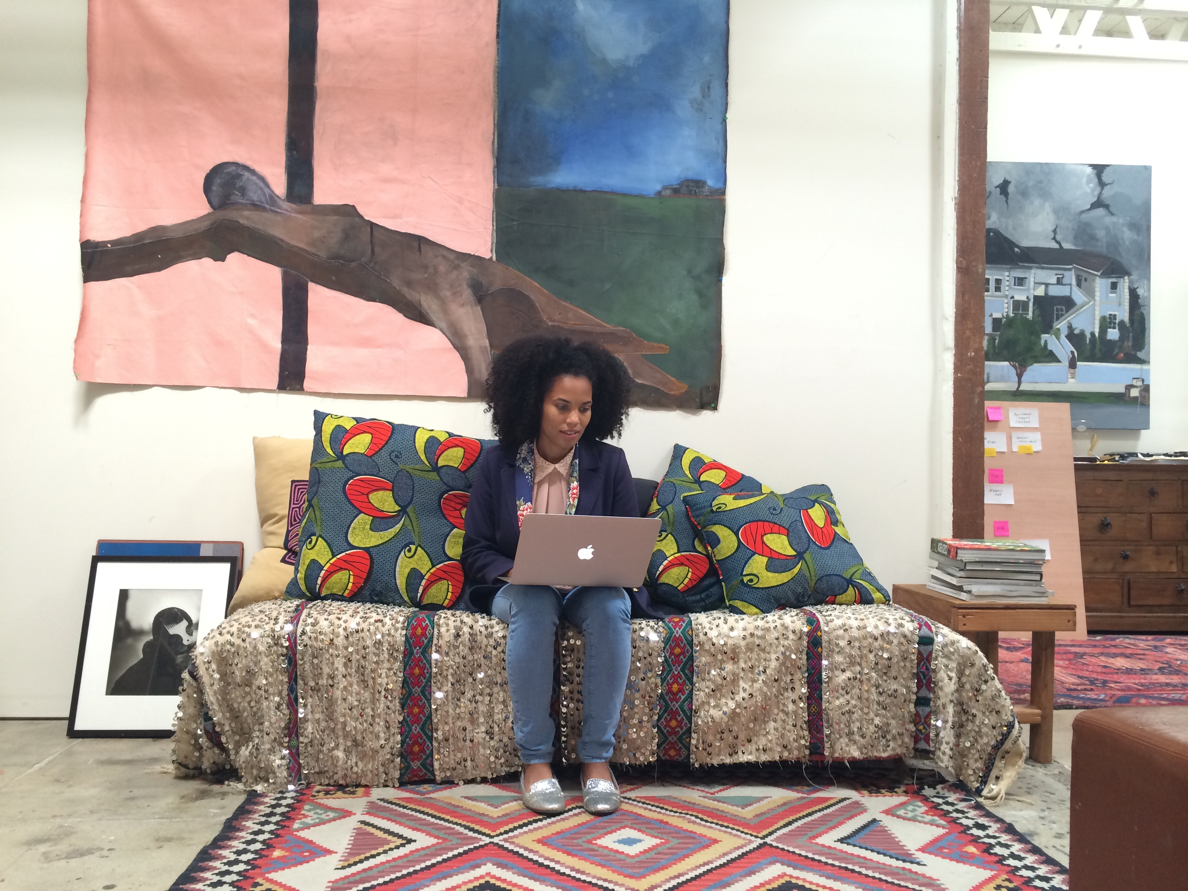 A person is sitting on a decorated sofa using a laptop in a colorful, art-filled room.