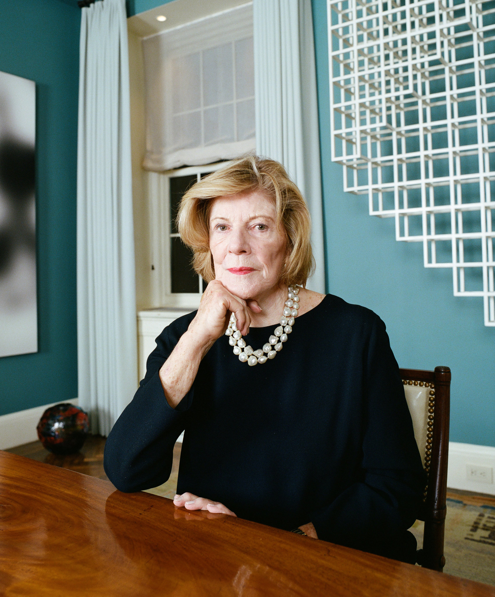 A woman in a dark dress sits at a wooden table with her face obscured and wears a pearl necklace.