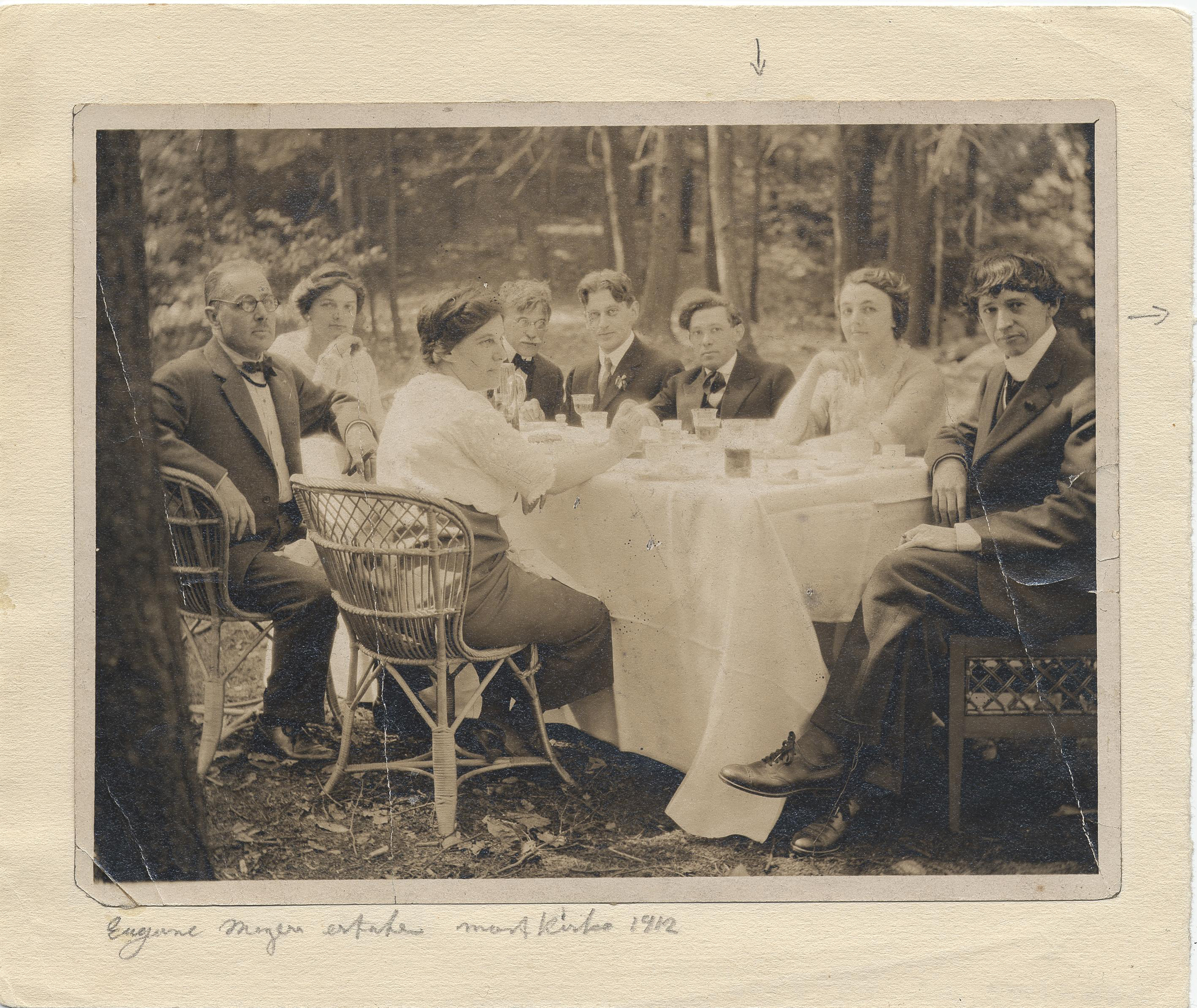 A group of people sit around a table outdoors, enjoying food and drinks in a wooded setting.