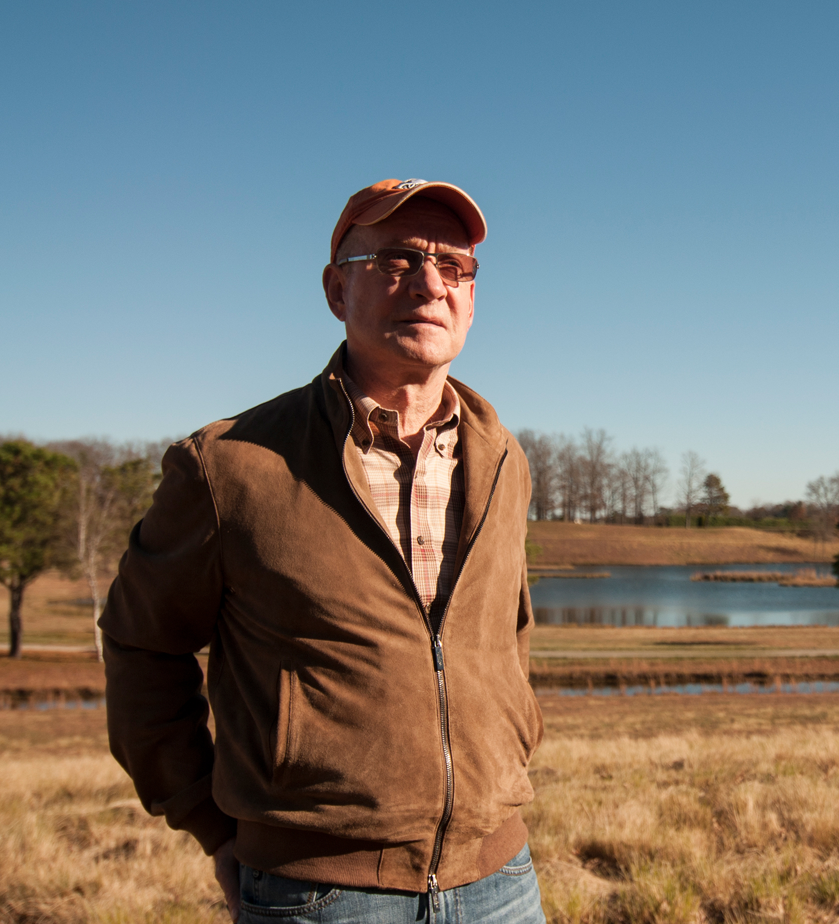 A person in a brown jacket and orange cap stands outdoors near a pond and grassy field.
