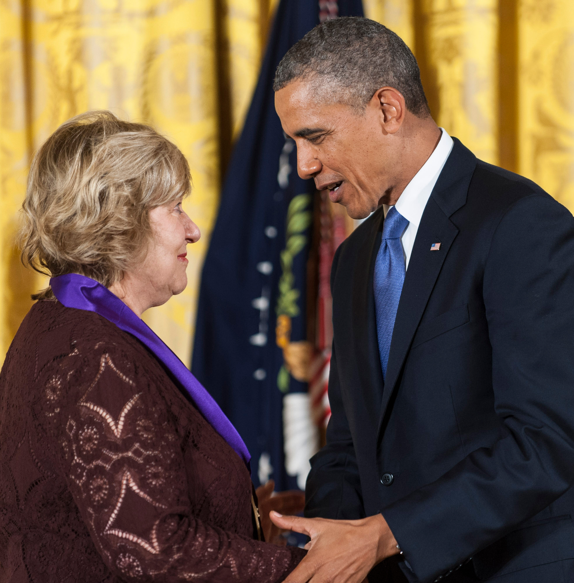 A man in a suit hands an award to a woman wearing a purple ribbon in a formal setting.