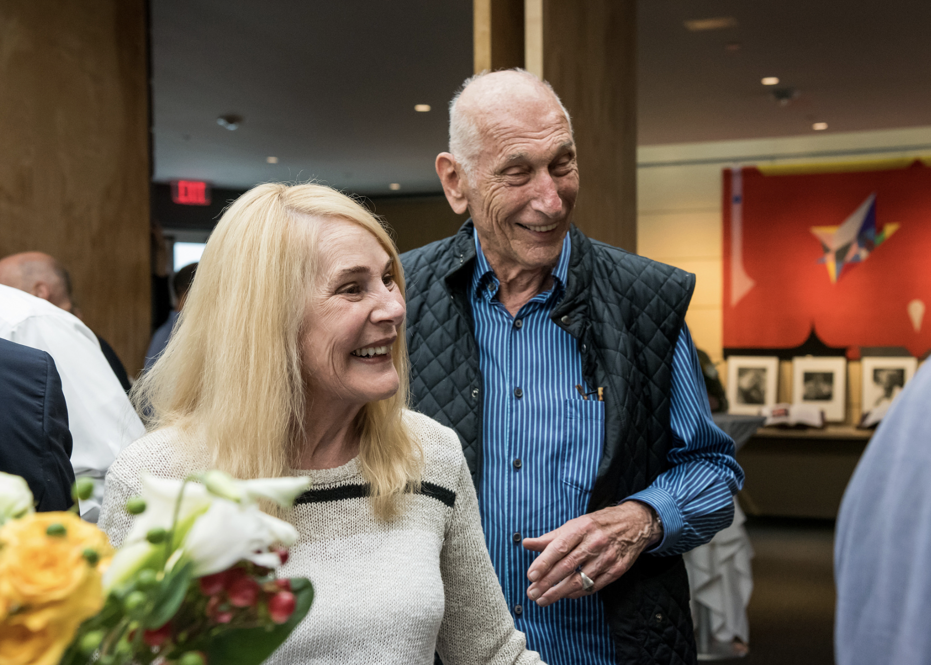 Two people stand indoors, one holding flowers, with blurred faces and art visible in the background.