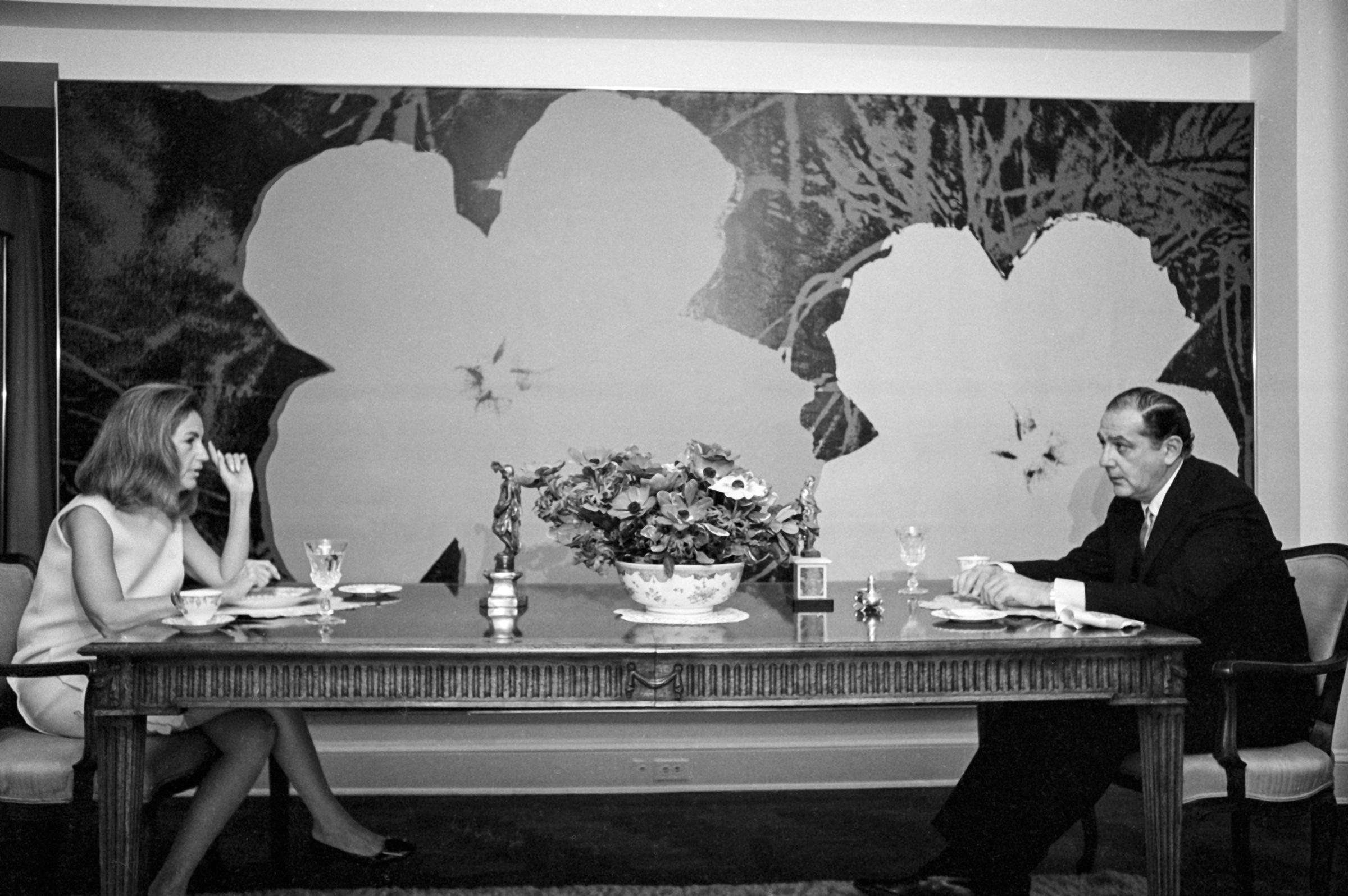 A man and woman dine at a table with a large floral artwork on the wall behind them.