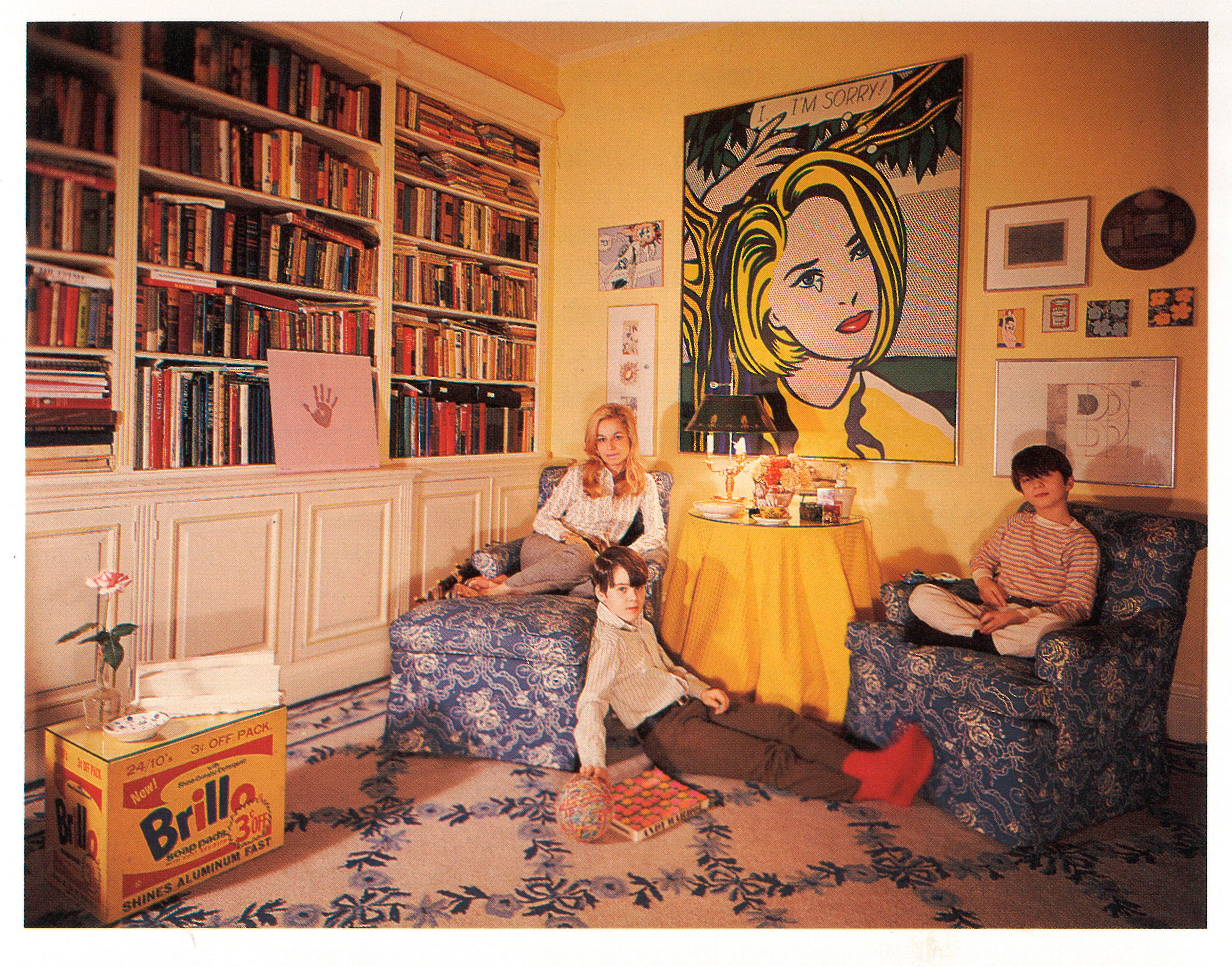 Three people relax in a cozy room with bookshelves, artwork, and patterned furniture.