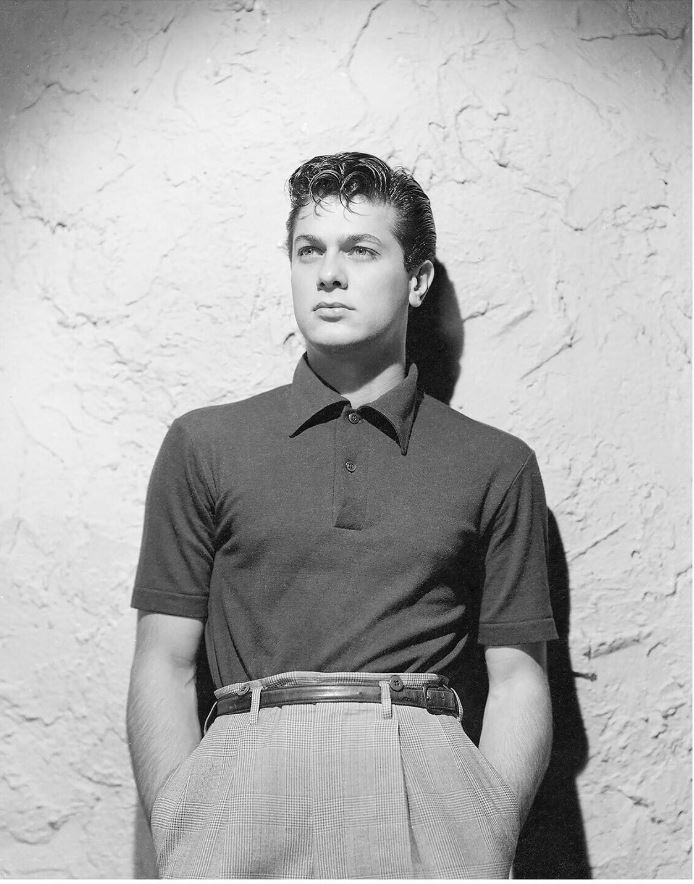 A man in a polo shirt and high-waisted pants stands against a textured wall, face blurred.