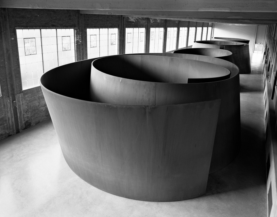 Large curved steel sculptures form spiral shapes inside a spacious industrial gallery.