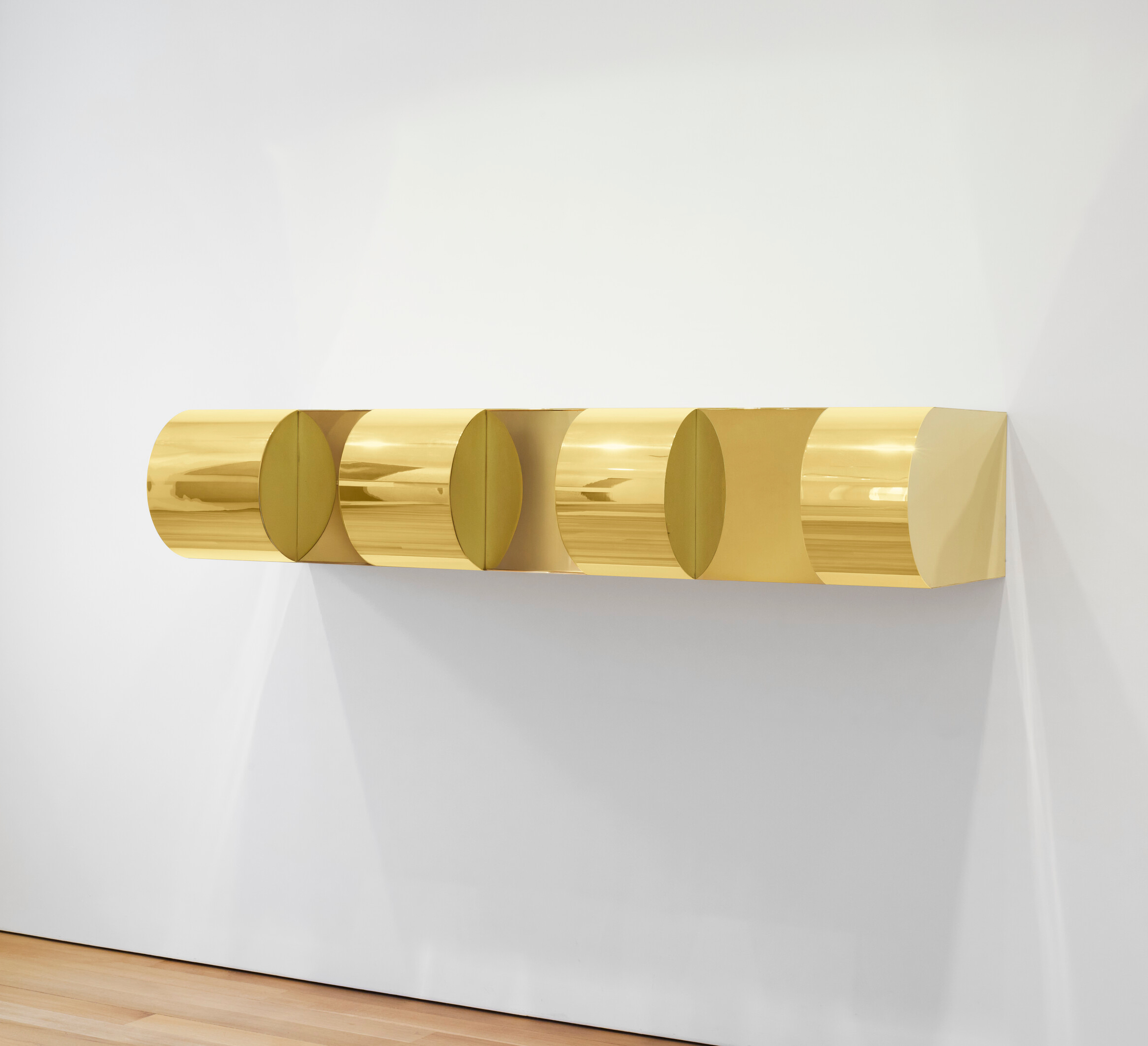 A shiny gold, cylindrical wall sculpture with five distinct segments mounted on a white wall.