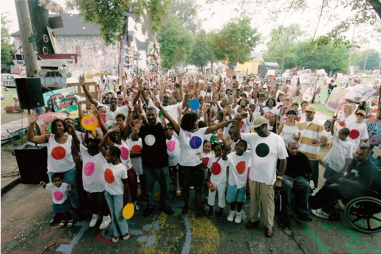 A large group of people wearing shirts with colorful circles gathers outdoors, raising their hands.