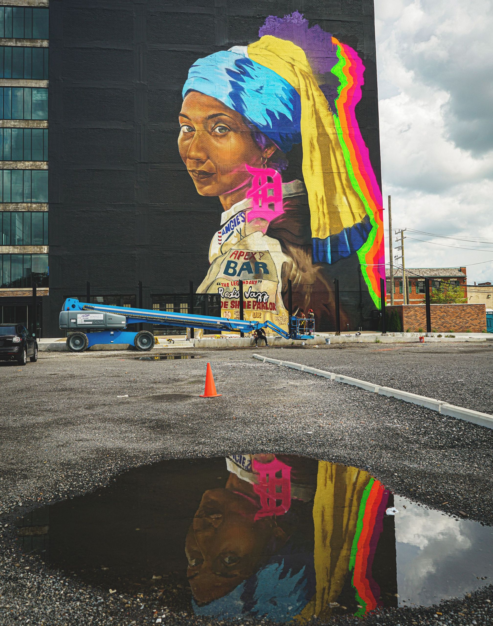 A colorful mural inspired by "Girl with a Pearl Earring" is reflected in a puddle in a parking lot.