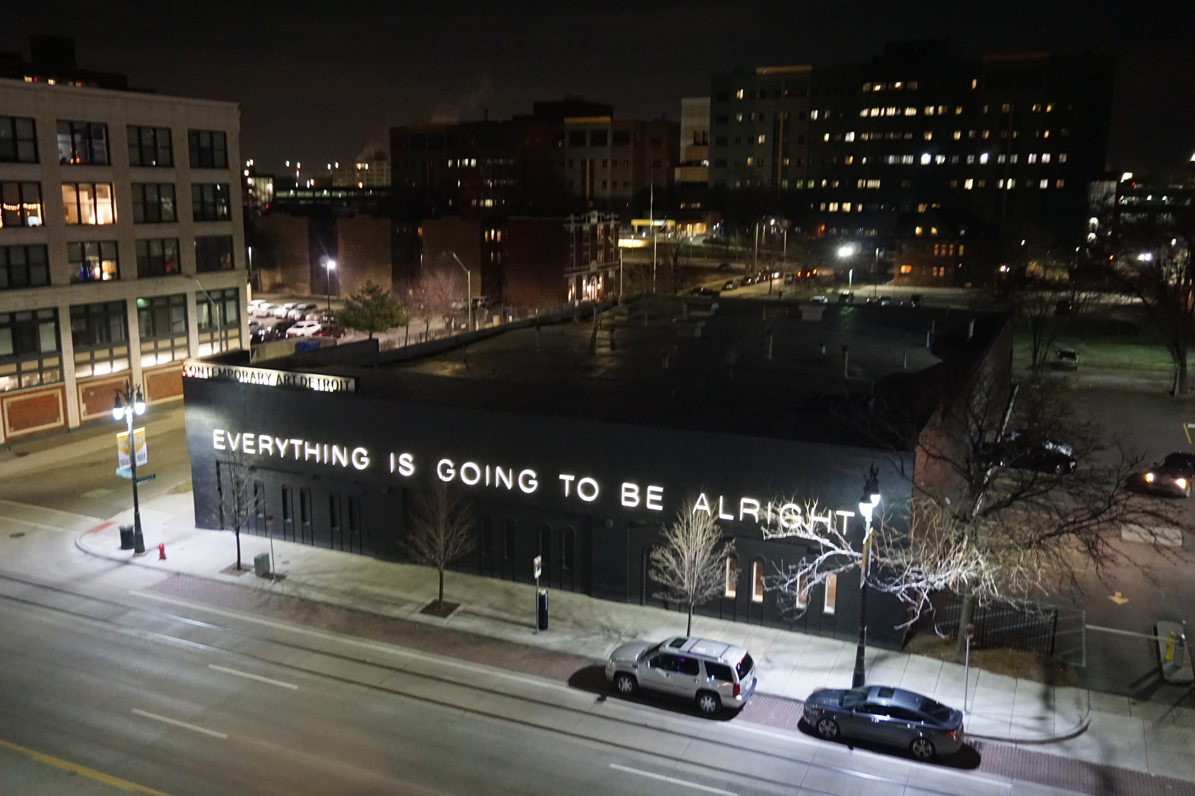 A building displays a bright sign reading "EVERYTHING IS GOING TO BE ALRIGHT" at night.