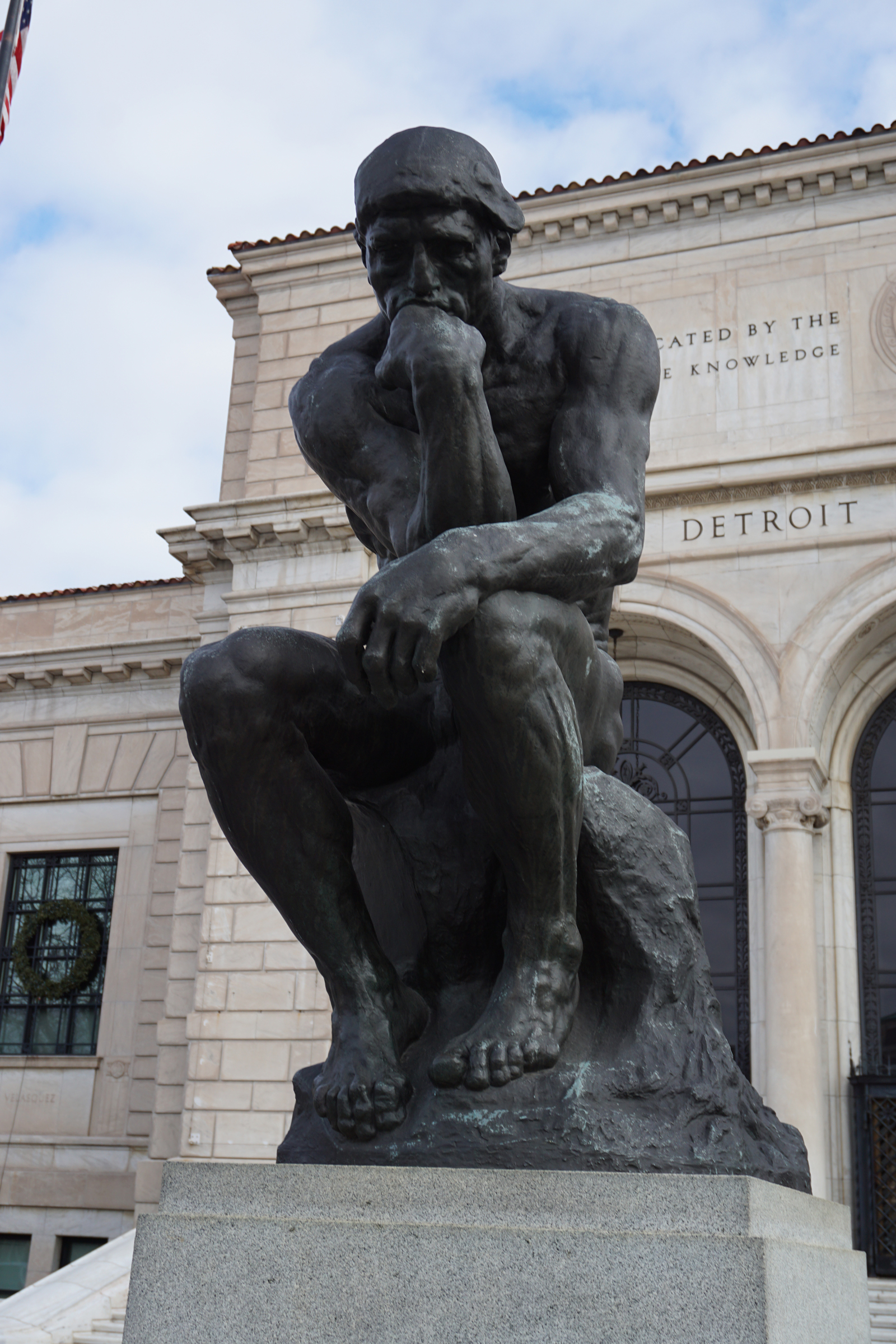 A bronze statue of a man deep in thought sits in front of a large stone building.