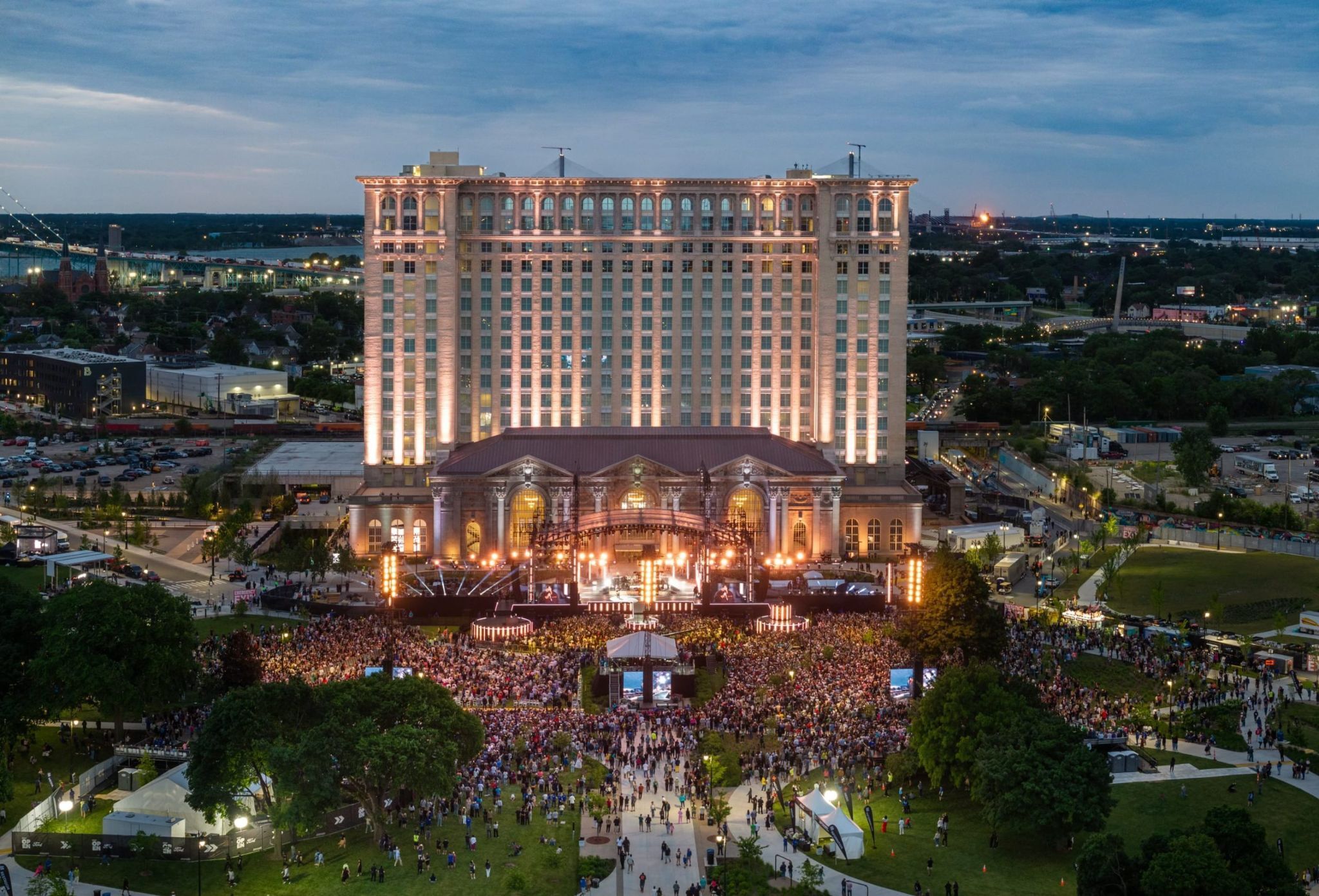 A large crowd gathers at an outdoor concert in front of an illuminated historic building at dusk.