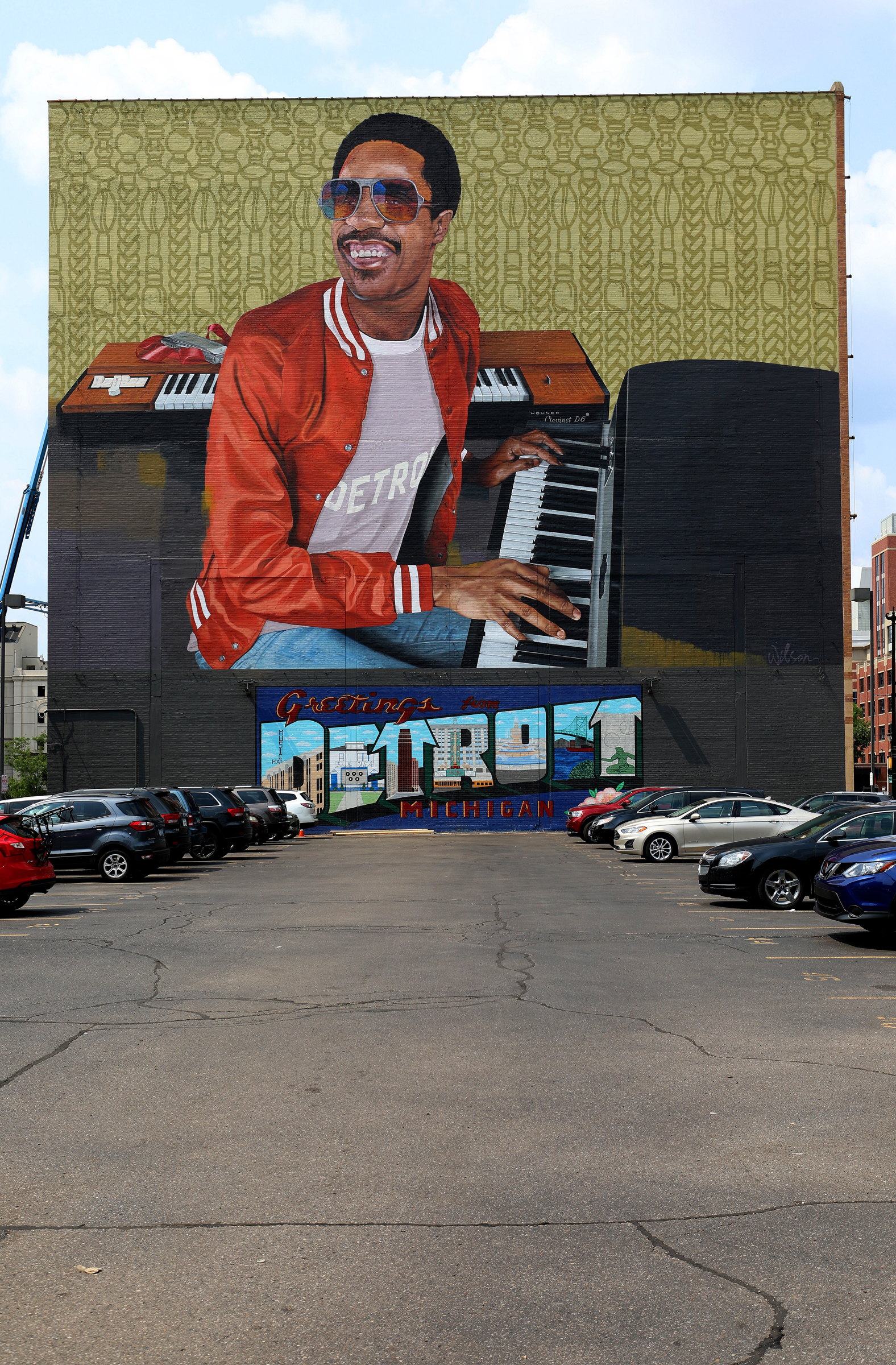 A mural of a man playing a keyboard is painted on a building above "Greetings from Detroit."