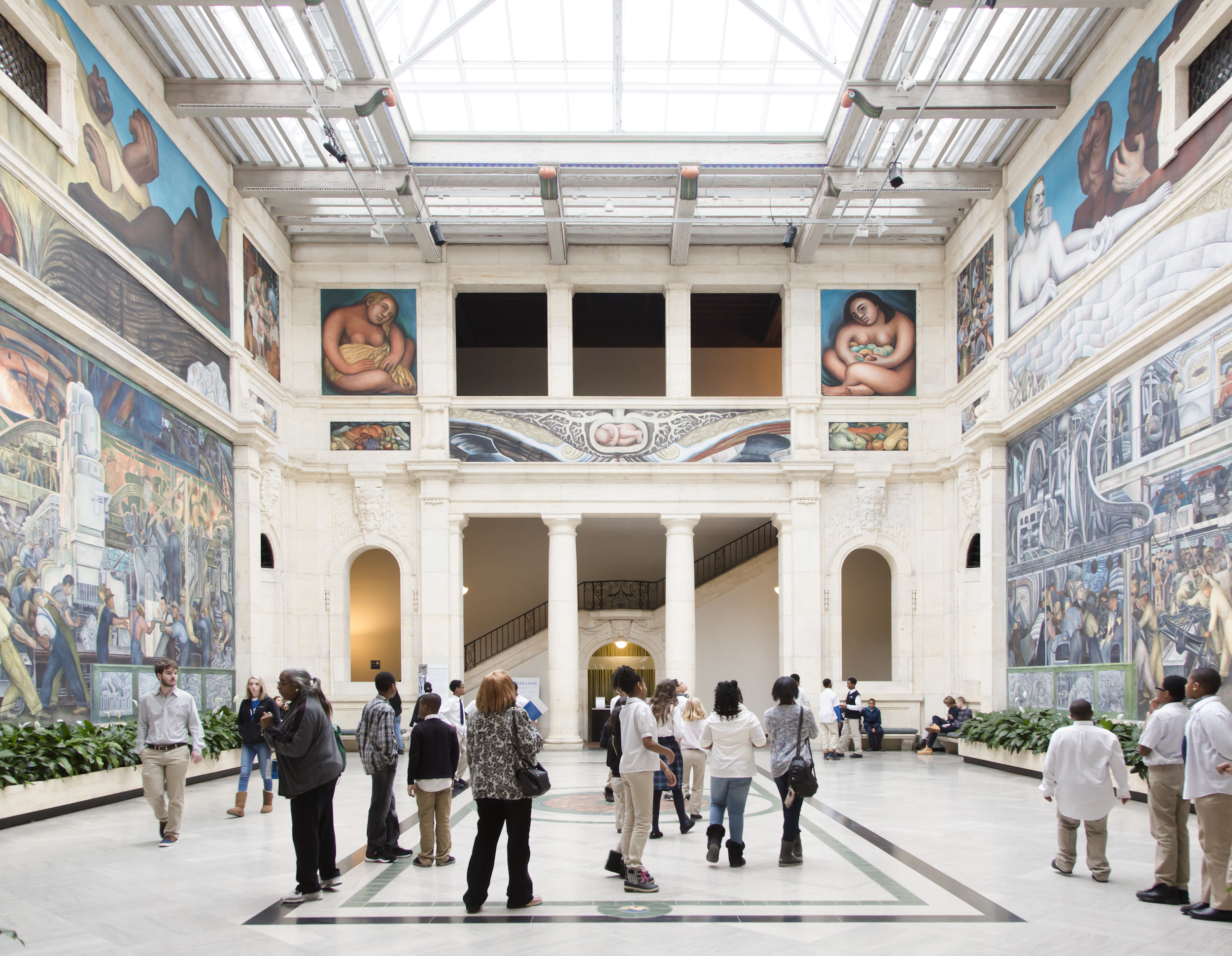 A group of people observe large murals in a spacious, bright art museum atrium.