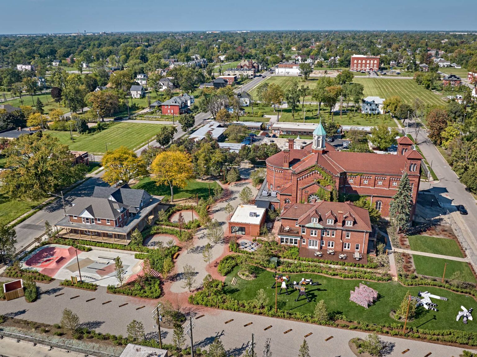 An aerial view of a neighborhood featuring a large brick building, homes, green spaces, and a skate park.