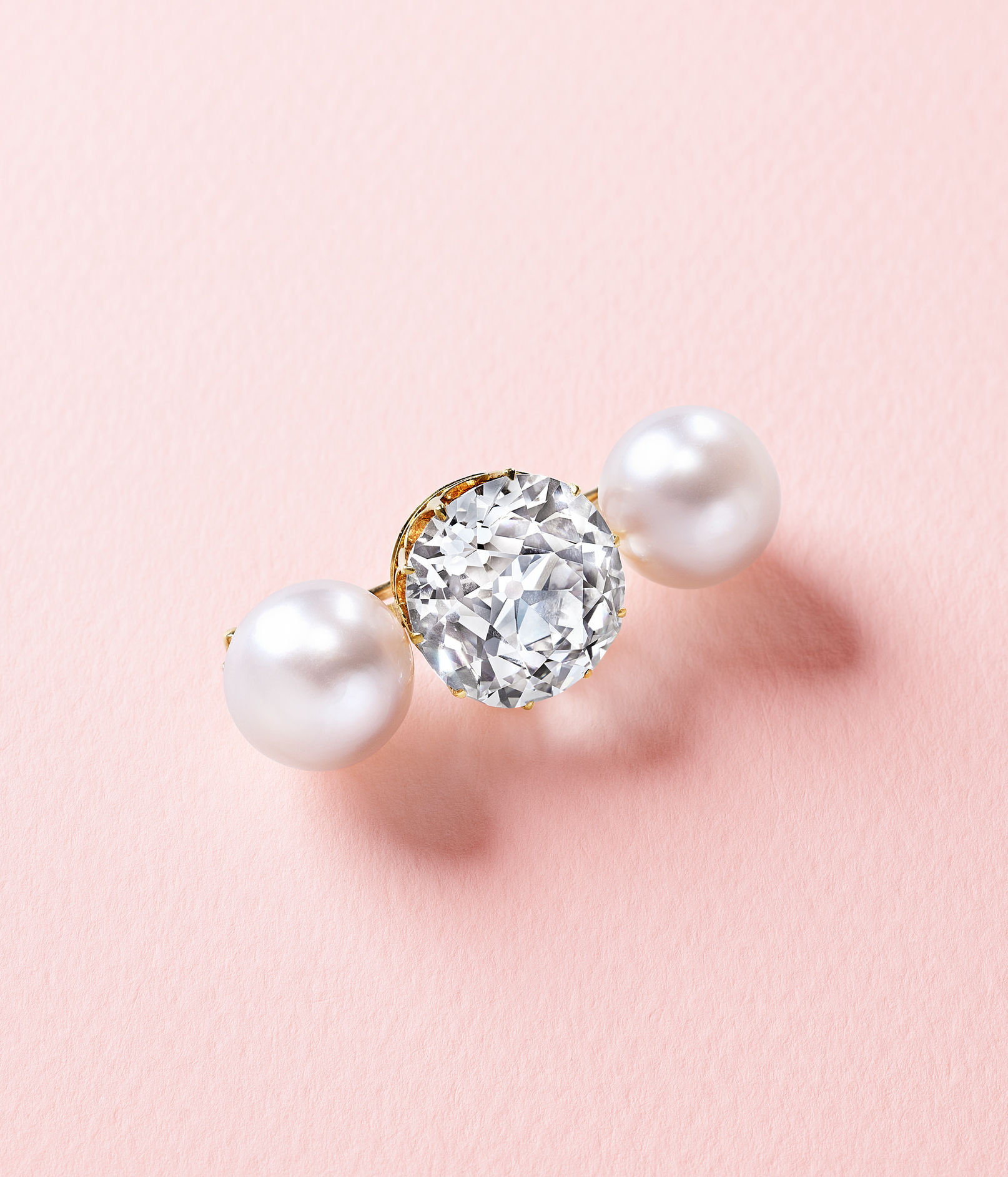 A gold ring with a large diamond in the center and a pearl on each side on a pink background.