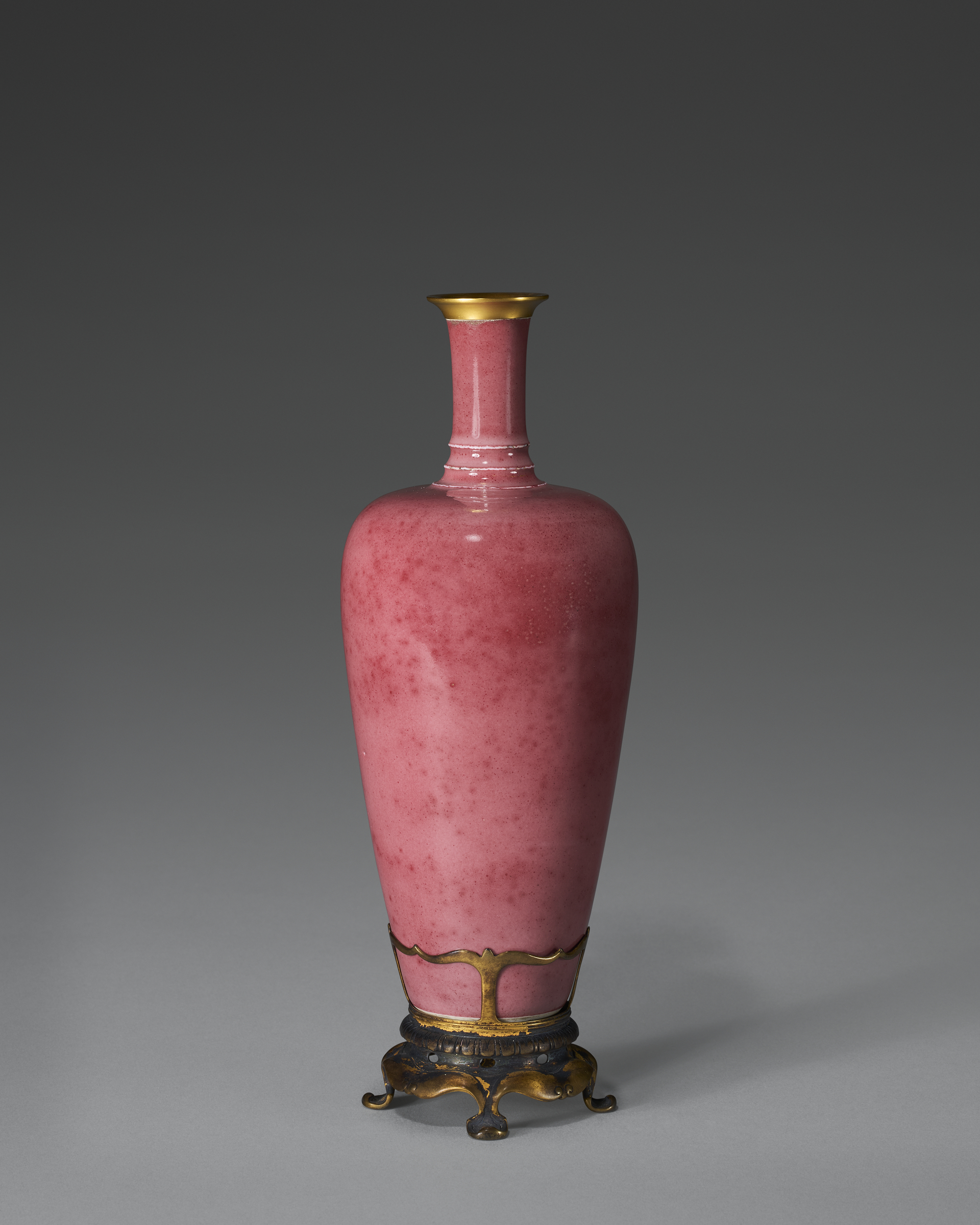 A tall pink ceramic vase with gold rim and ornate gold metal base against a gray background.