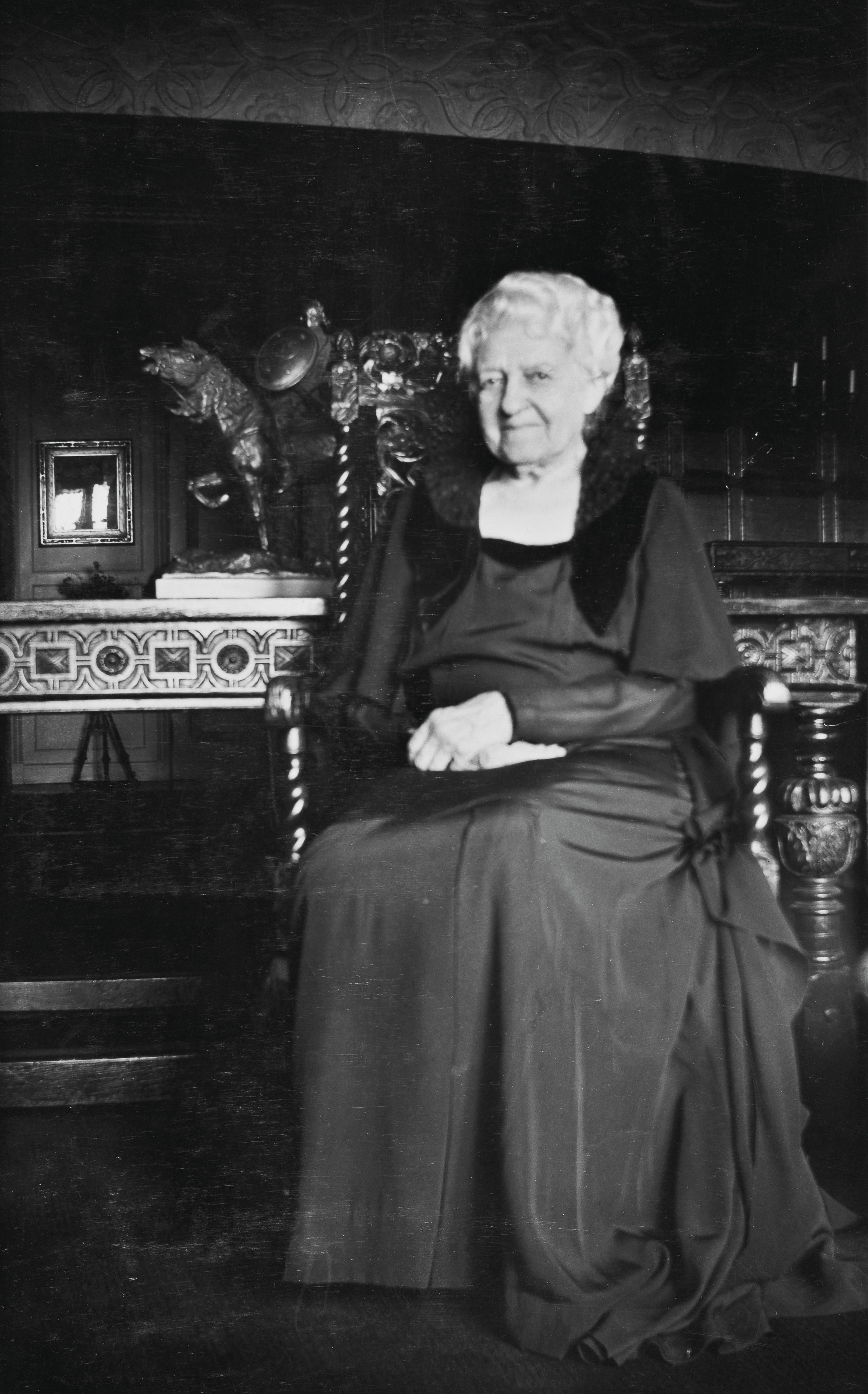 An elderly woman in a long dress sits on an ornate chair in a dimly lit room.
