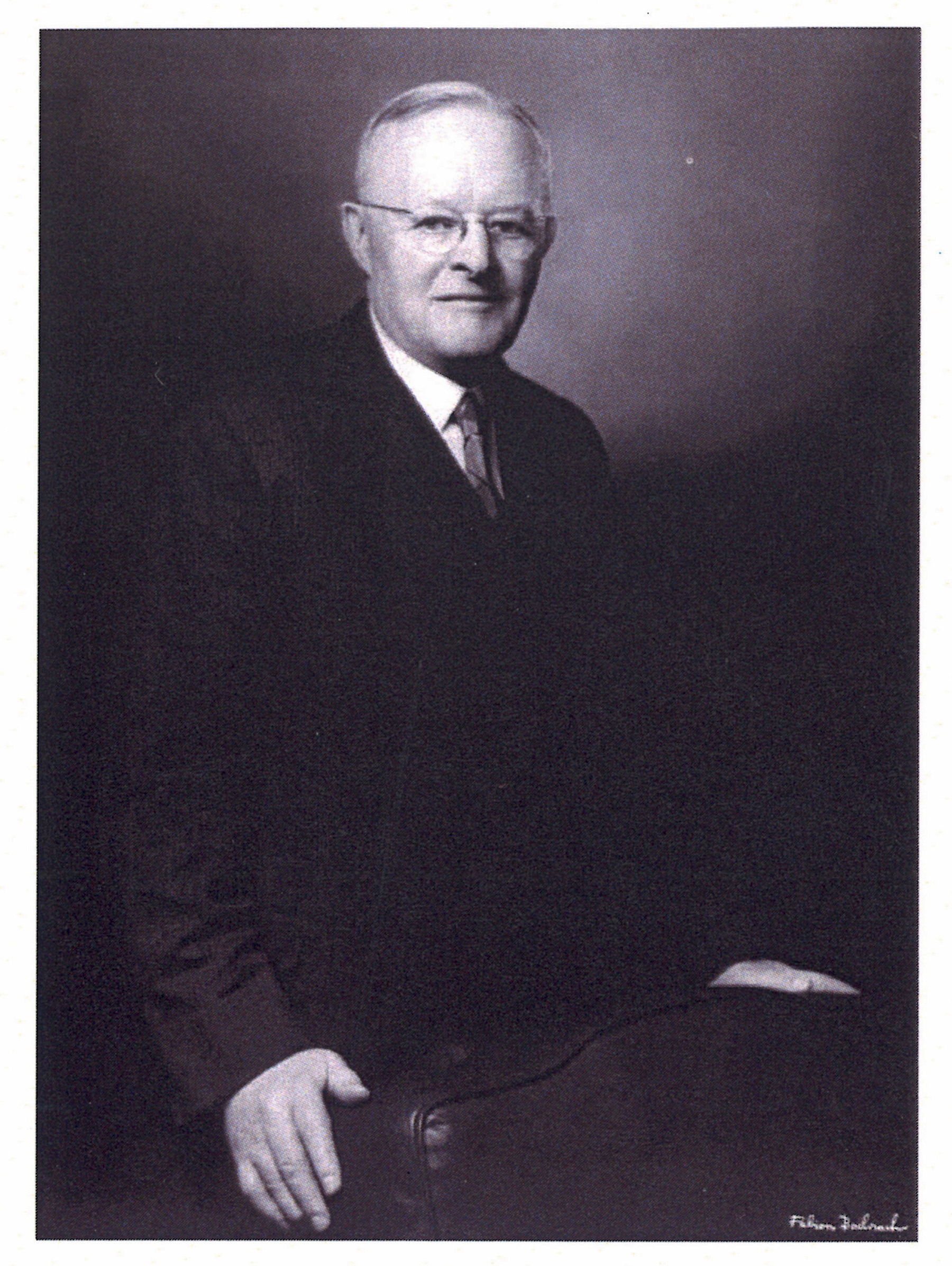An elderly man in a suit and glasses stands with one hand on a chair in a studio portrait.