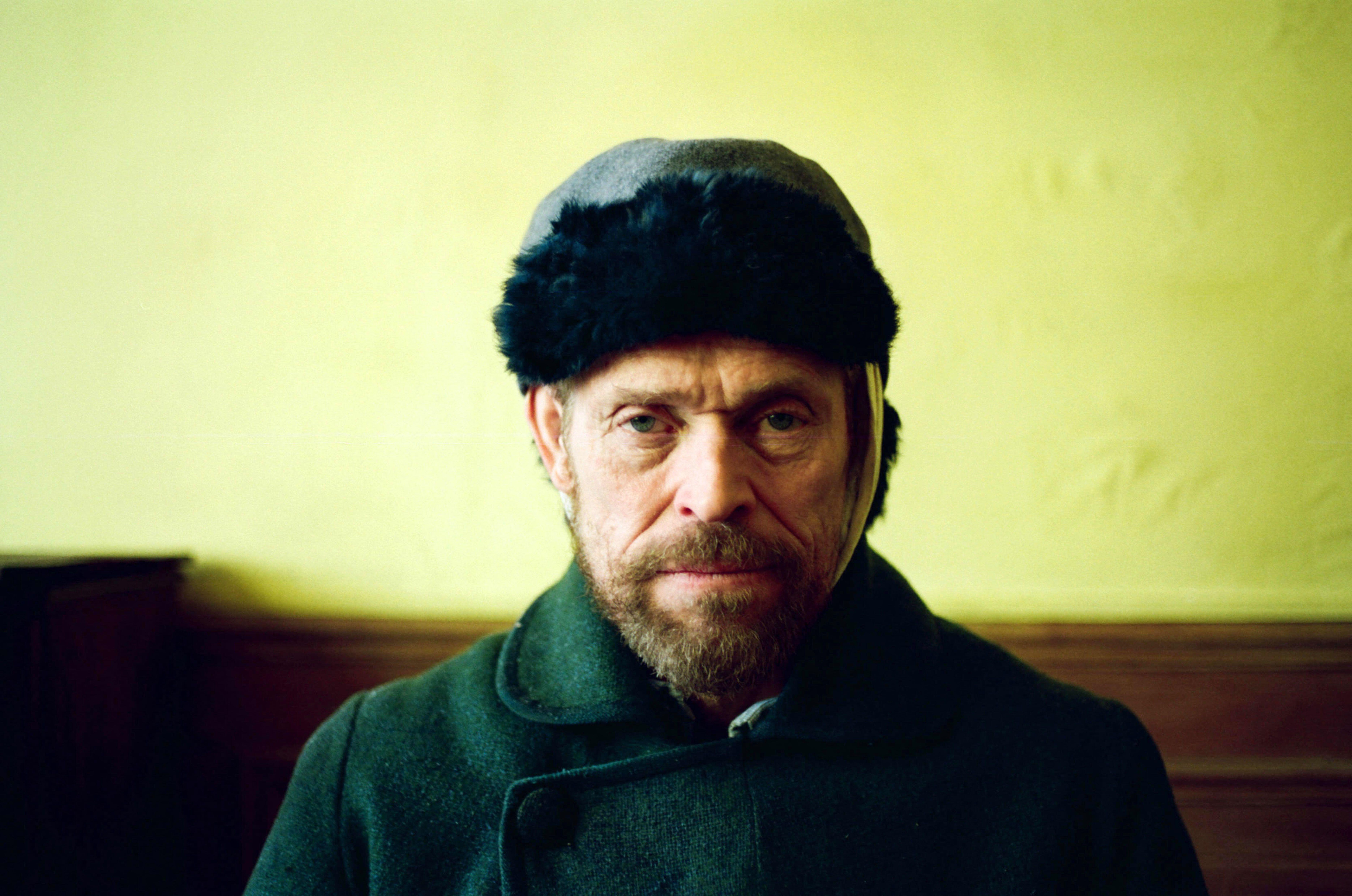 A man with a beard wearing a dark coat and fur hat sits against a yellow wall.